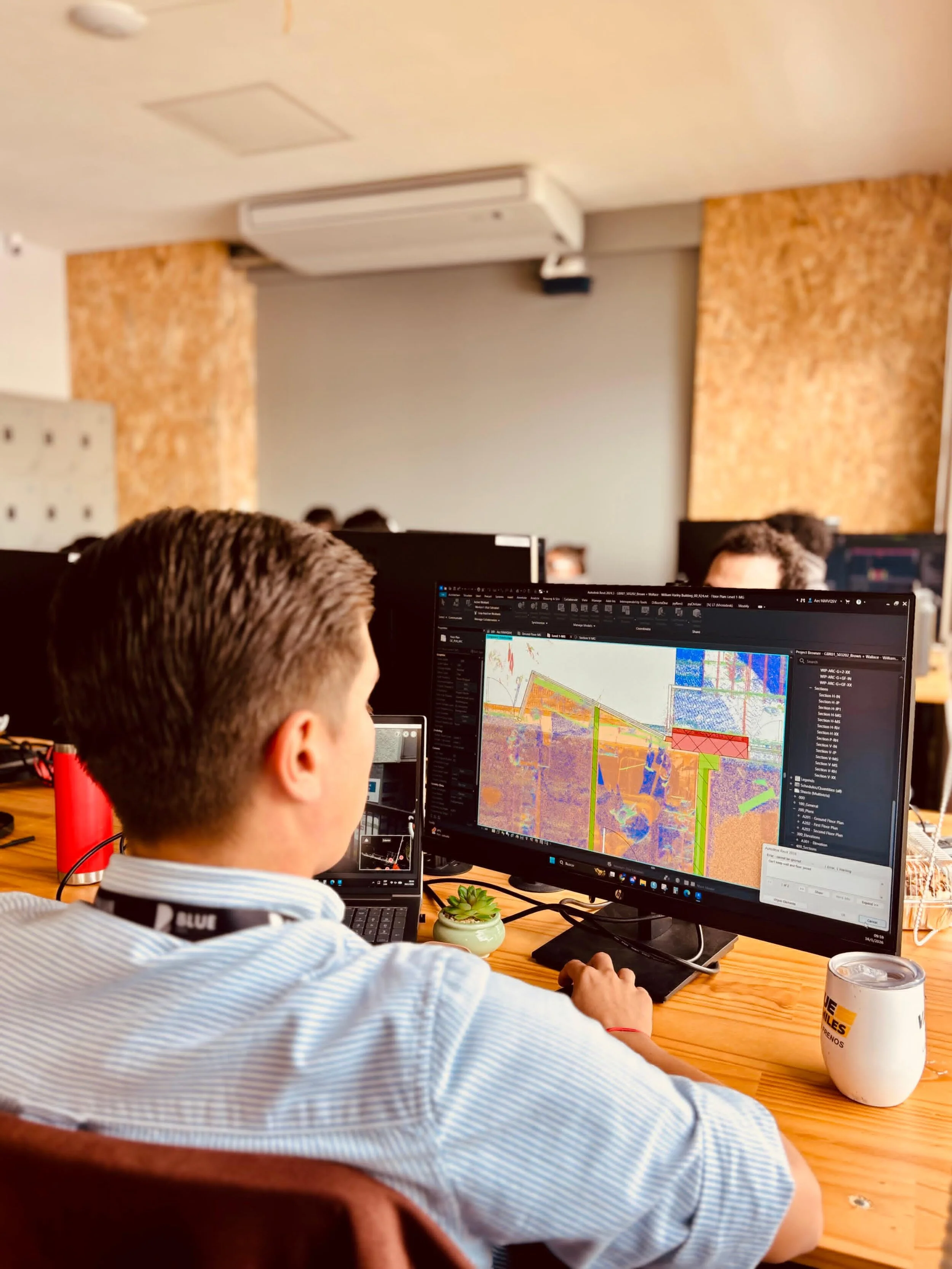 A man in a light blue striped shirt working on a desktop computer displaying colorful 3D modeling or mapping software, with a smaller monitor to the left, a succulent plant in a small pot, a red thermos, and a white coffee mug on a wooden table in a modern office or workspace.
