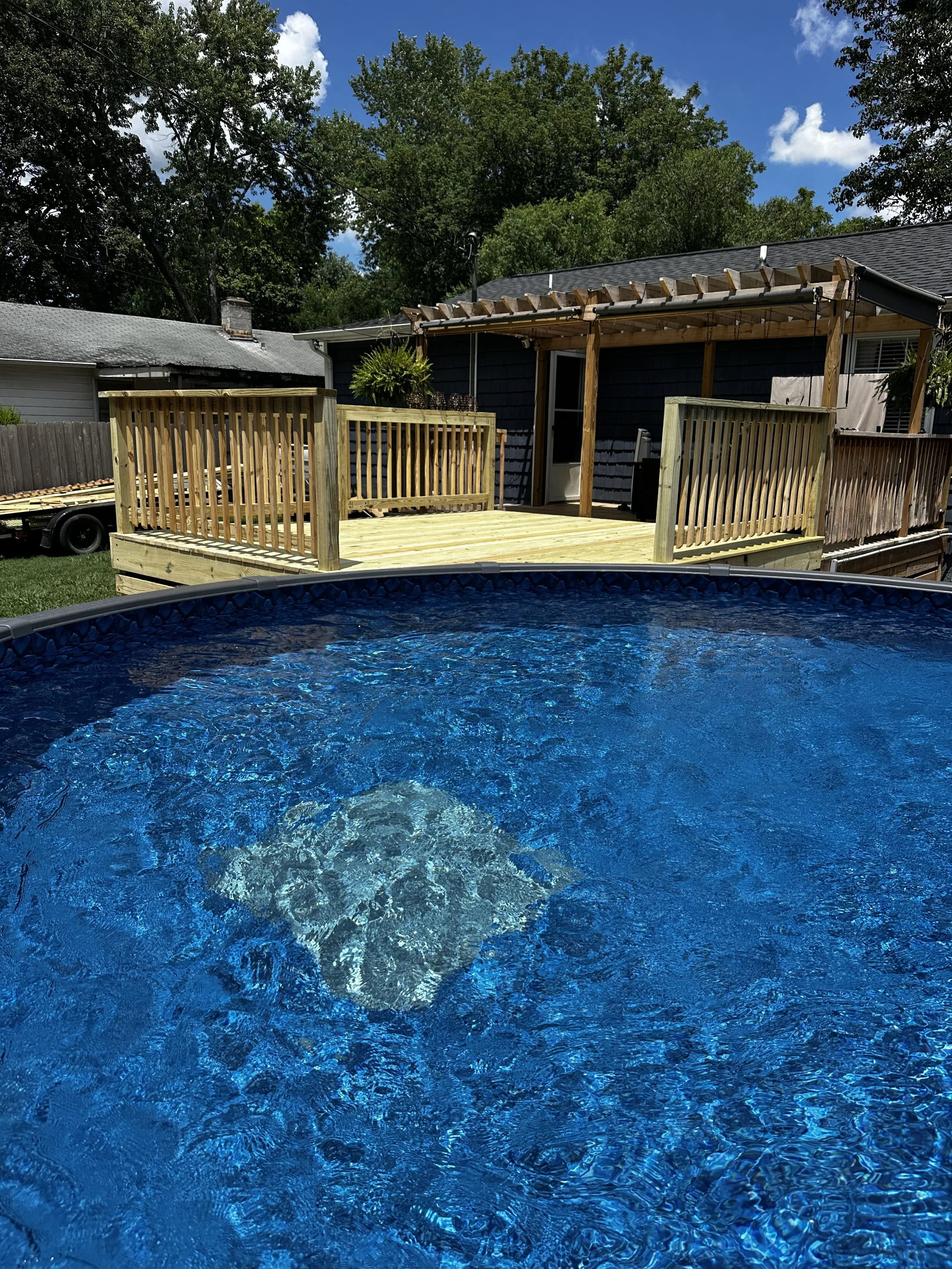 Backyard with a above ground swimming pool in the foreground and a wooden deck attached to a dark blue house in the background.