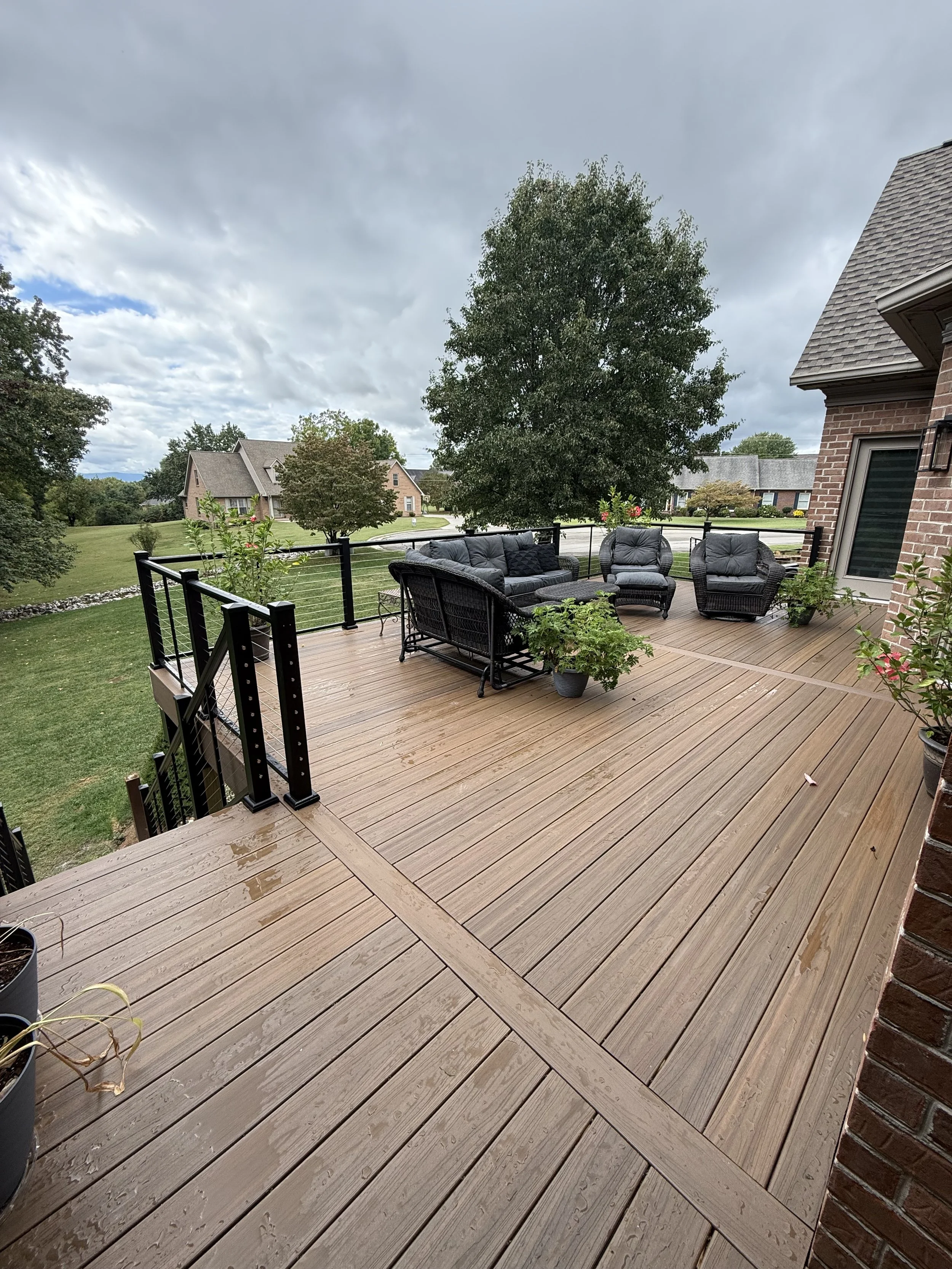 A wooden outdoor deck with black patio furniture including a sofa and chairs, potted plants, and a black metal railing, overlooking a green yard with trees and neighboring houses under a cloudy sky.