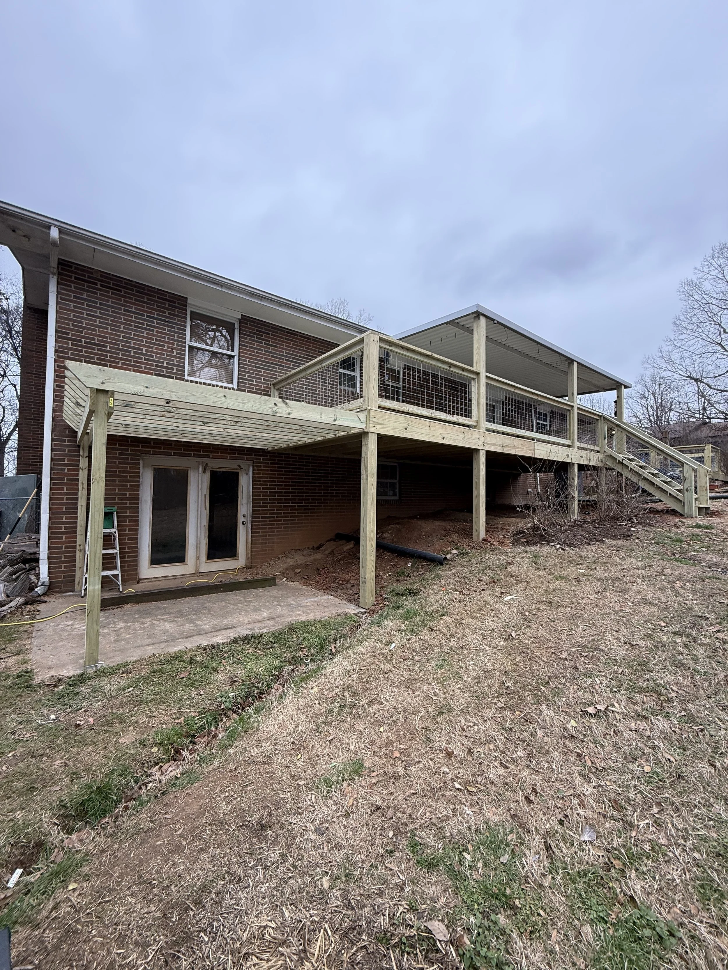 Newly constructed wooden deck and stairs attached to the back of a brick house with a sliding glass door leading to the backyard, on a cloudy day.