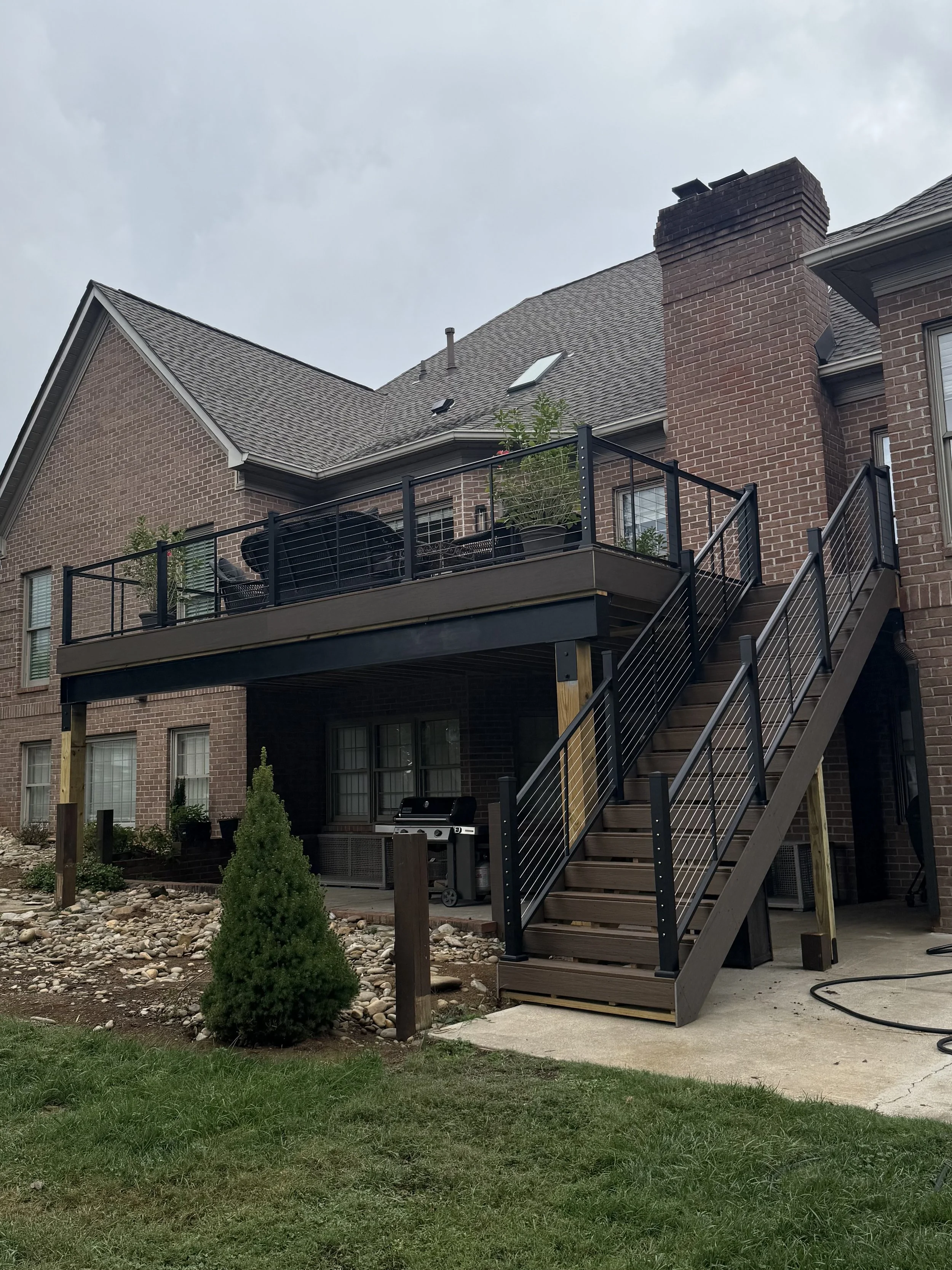 Backyard with a newly built wooden deck attached to a brick house, with stairs leading down to a grassy yard, a small evergreen bush, and a barbecue grill.
