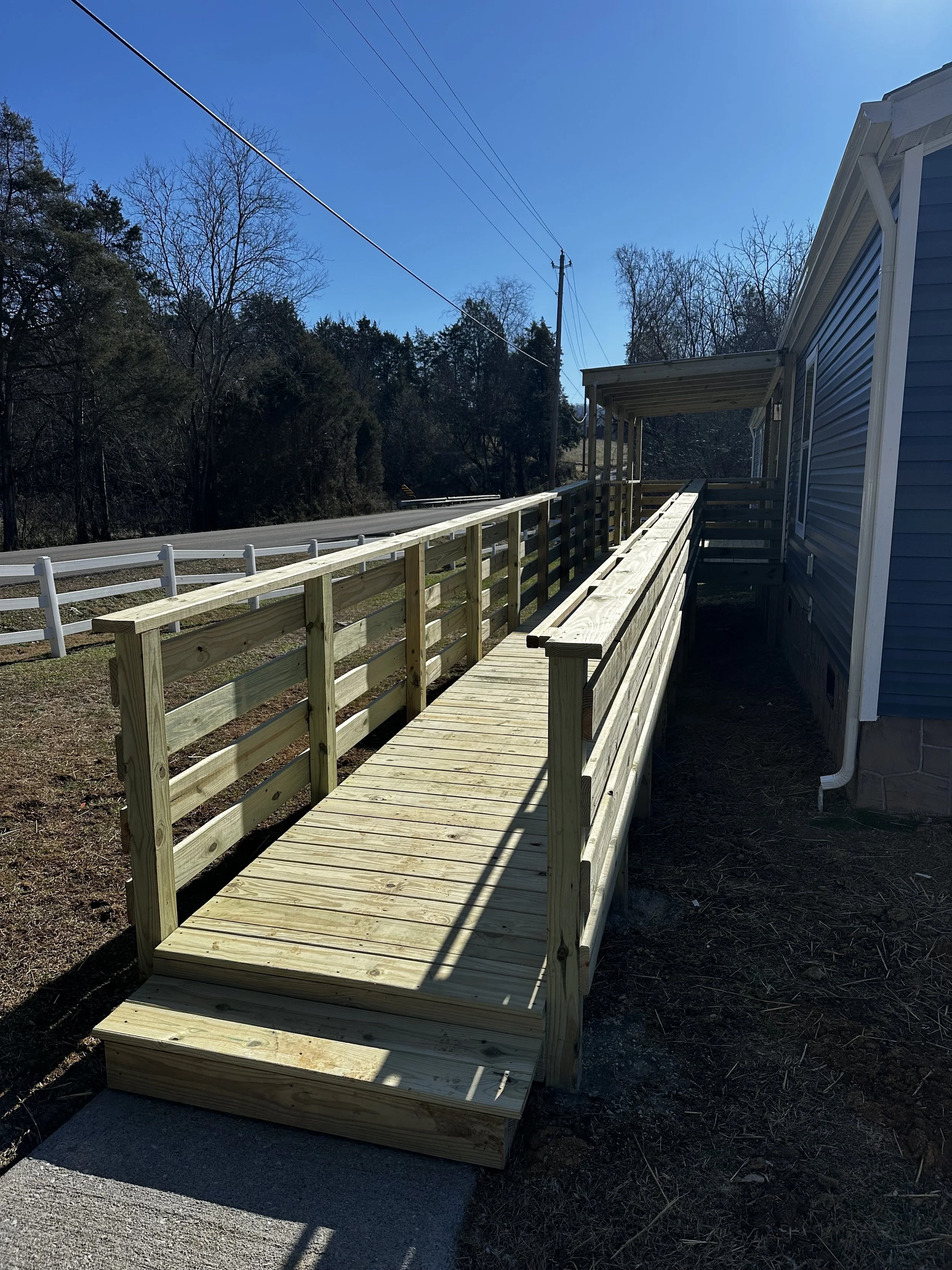 Wooden wheelchair accessible ramp attached to the side of a blue house, leading from a concrete sidewalk to the house entrance, with railings on both sides, under a bright blue sky.