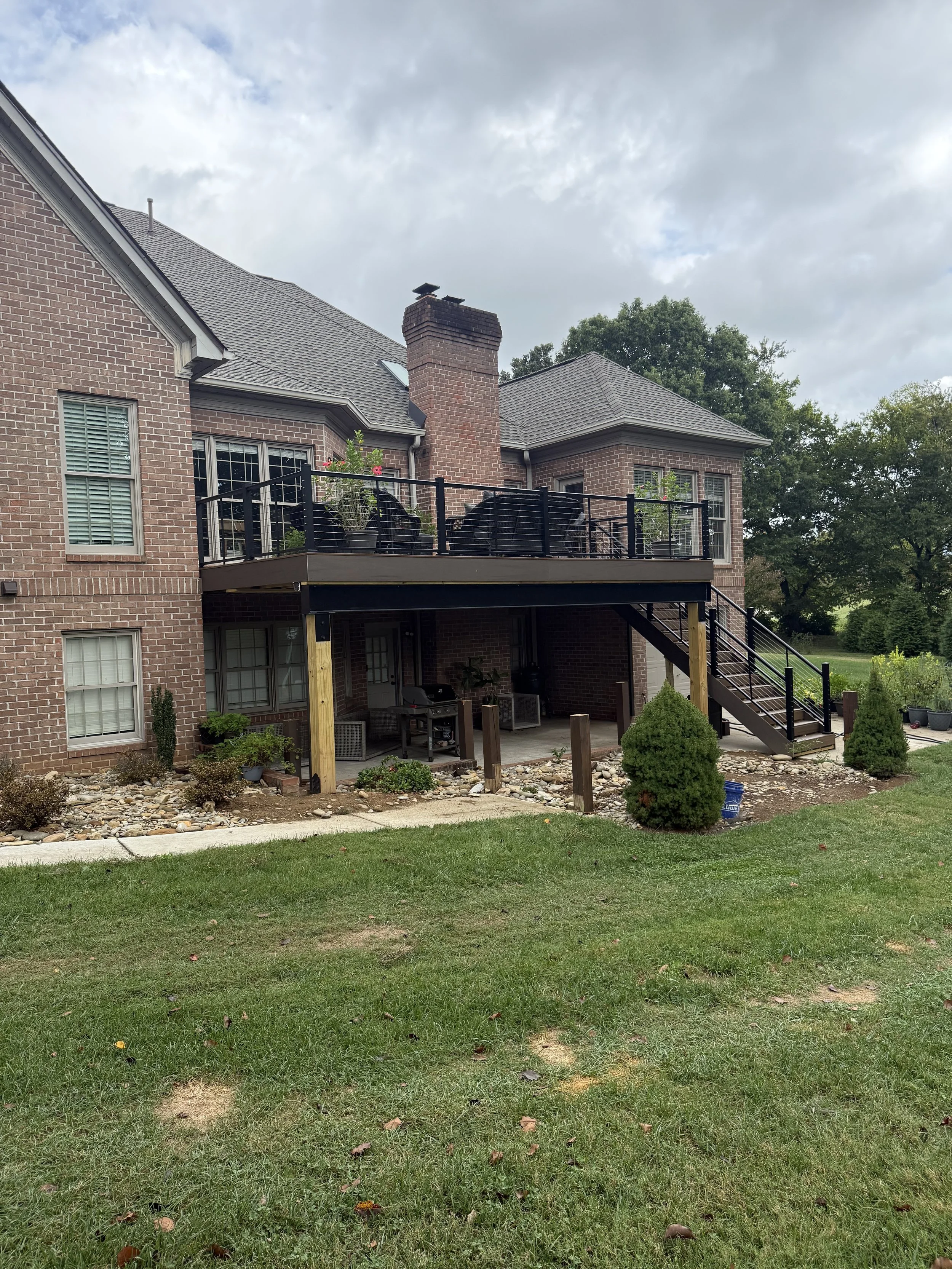 Backyard view of a brick house with a raised deck, outdoor stairs, patio furniture, plants, and small bushes, under a cloudy sky in Maryville,TN
