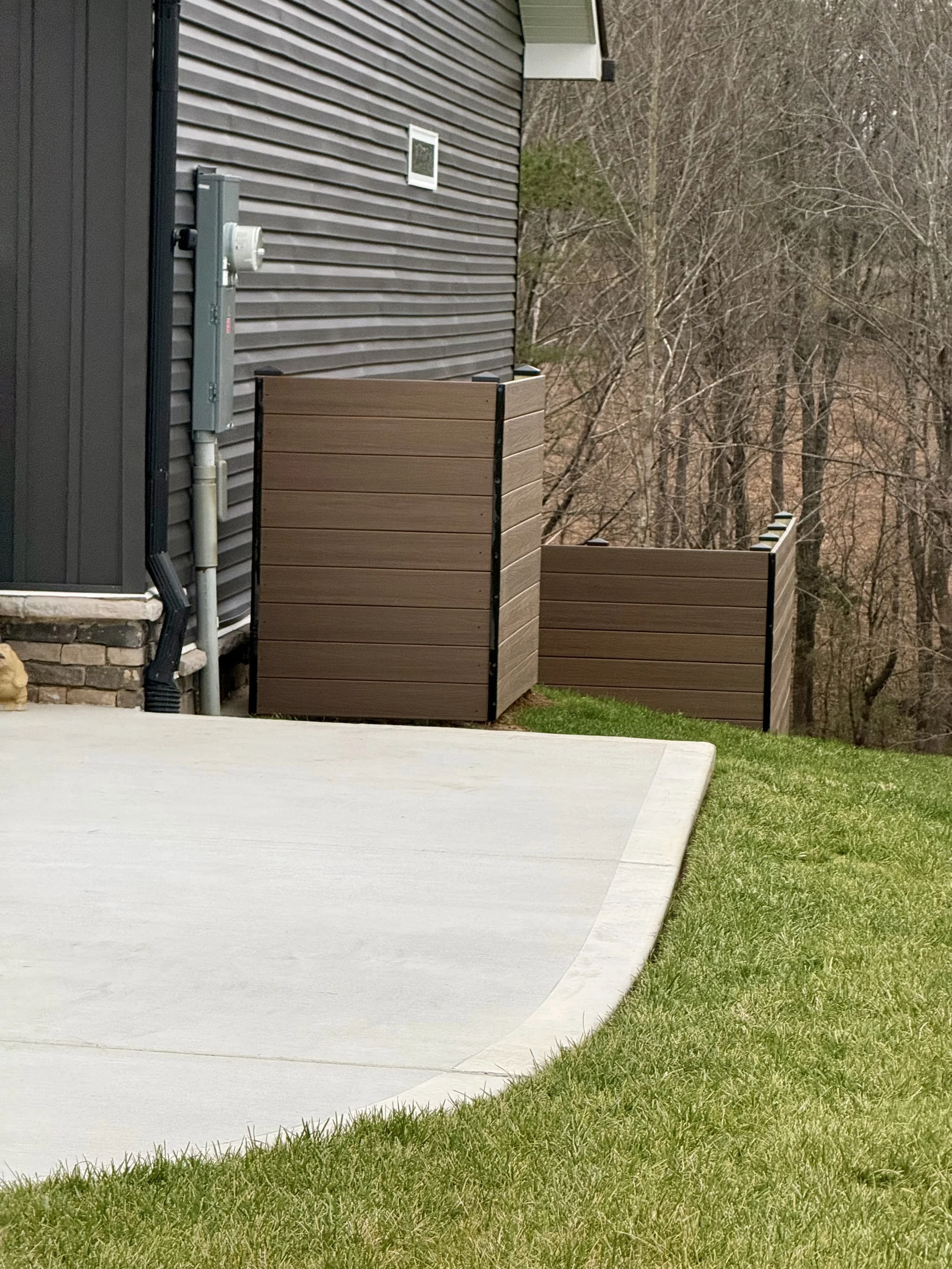 A section of a house exterior showing brown wooden privacy screens, a concrete patio, green grass, and a wooded background.
