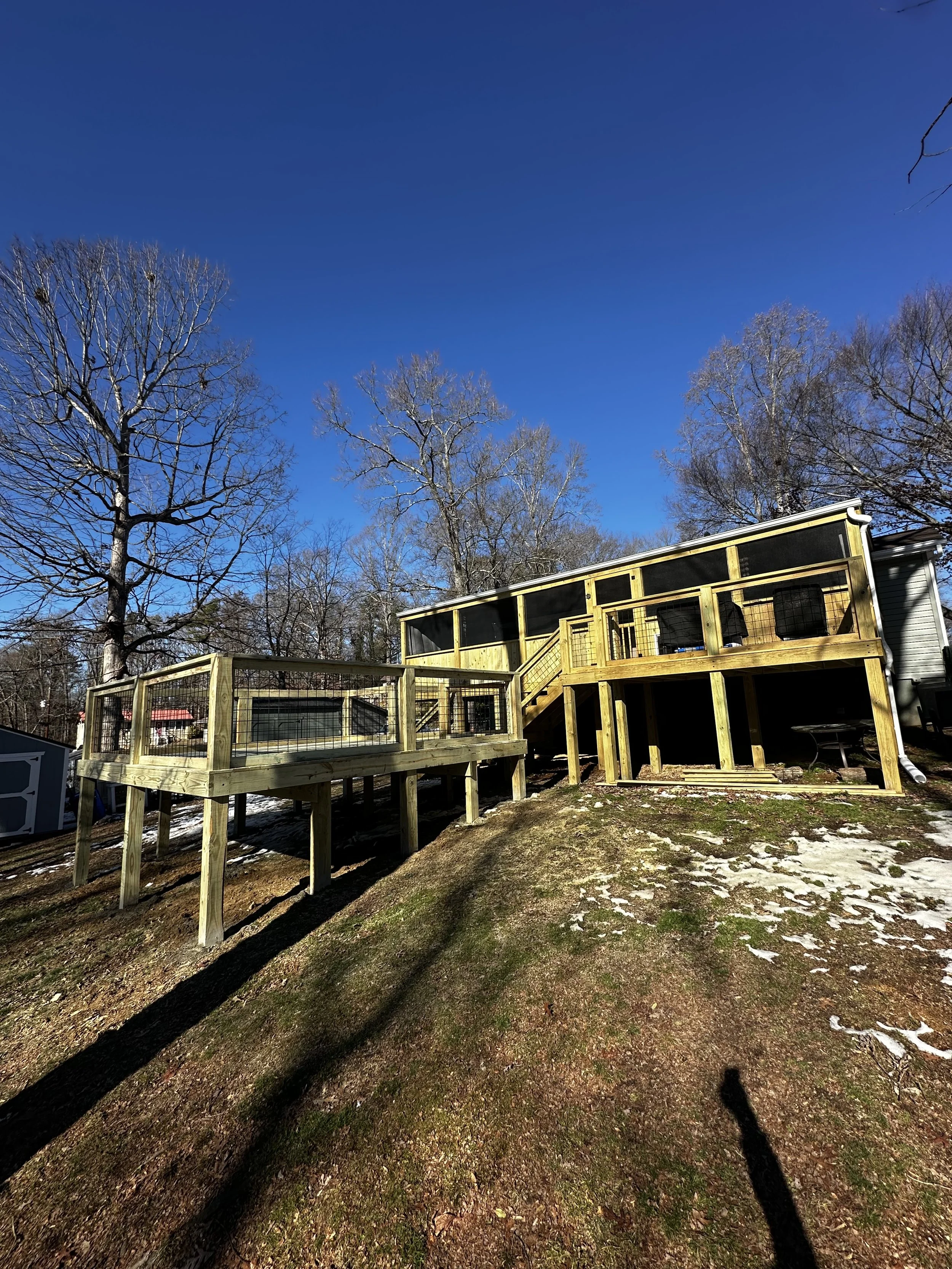 A large wooden deck with railing, stairs, and a screened enclosure attached to a house, set on a sloped yard with patches of snow, leafless trees in the background, and a bright blue sky.