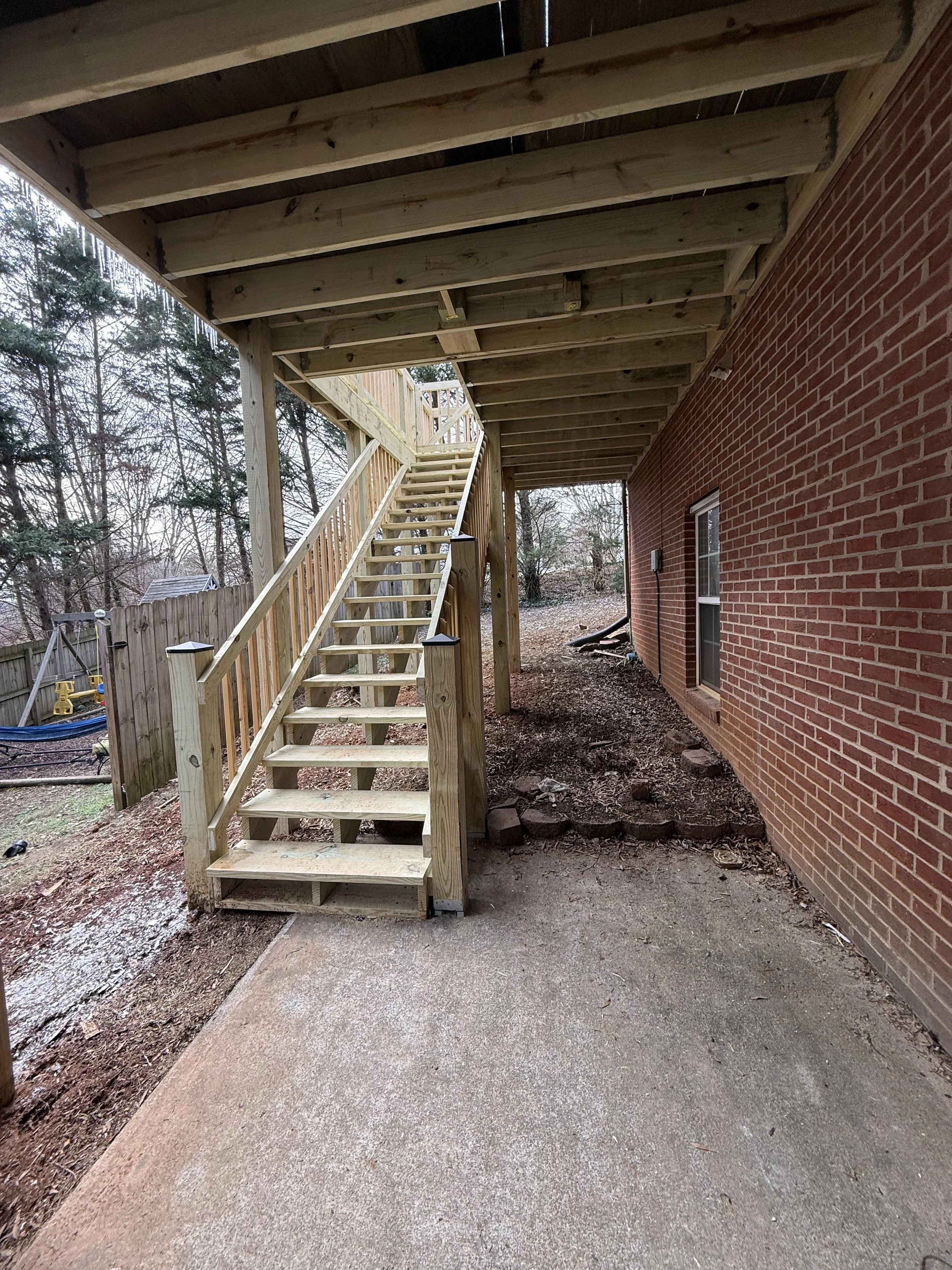 Wooden staircase leading up to a deck attached to the back of a brick house. The staircase has a handrail and is supported by posts. Surrounding area is bare ground with some bricks and construction materials.