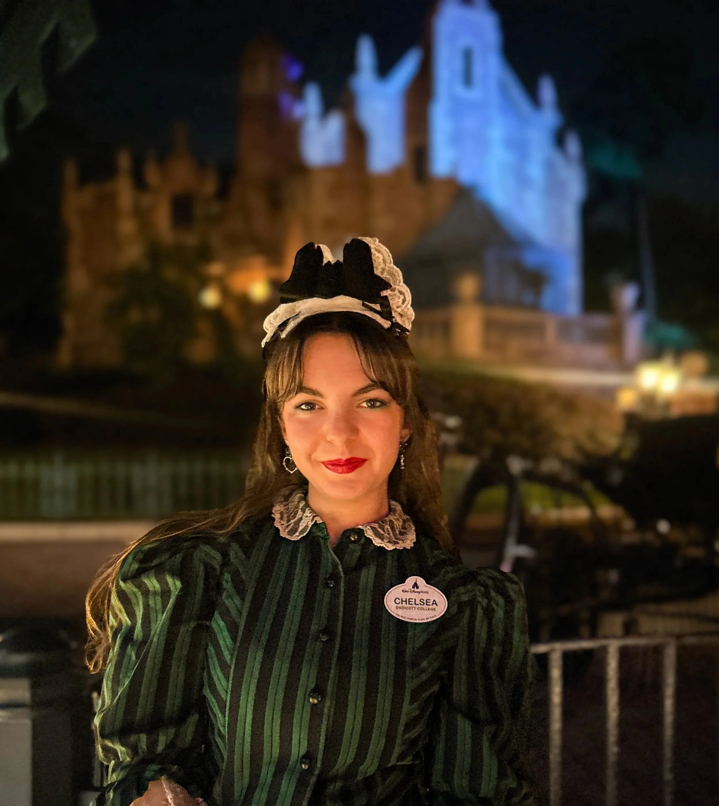Chelsea, in her Cast Member costume, stands outside the Haunted Mansion at night.
