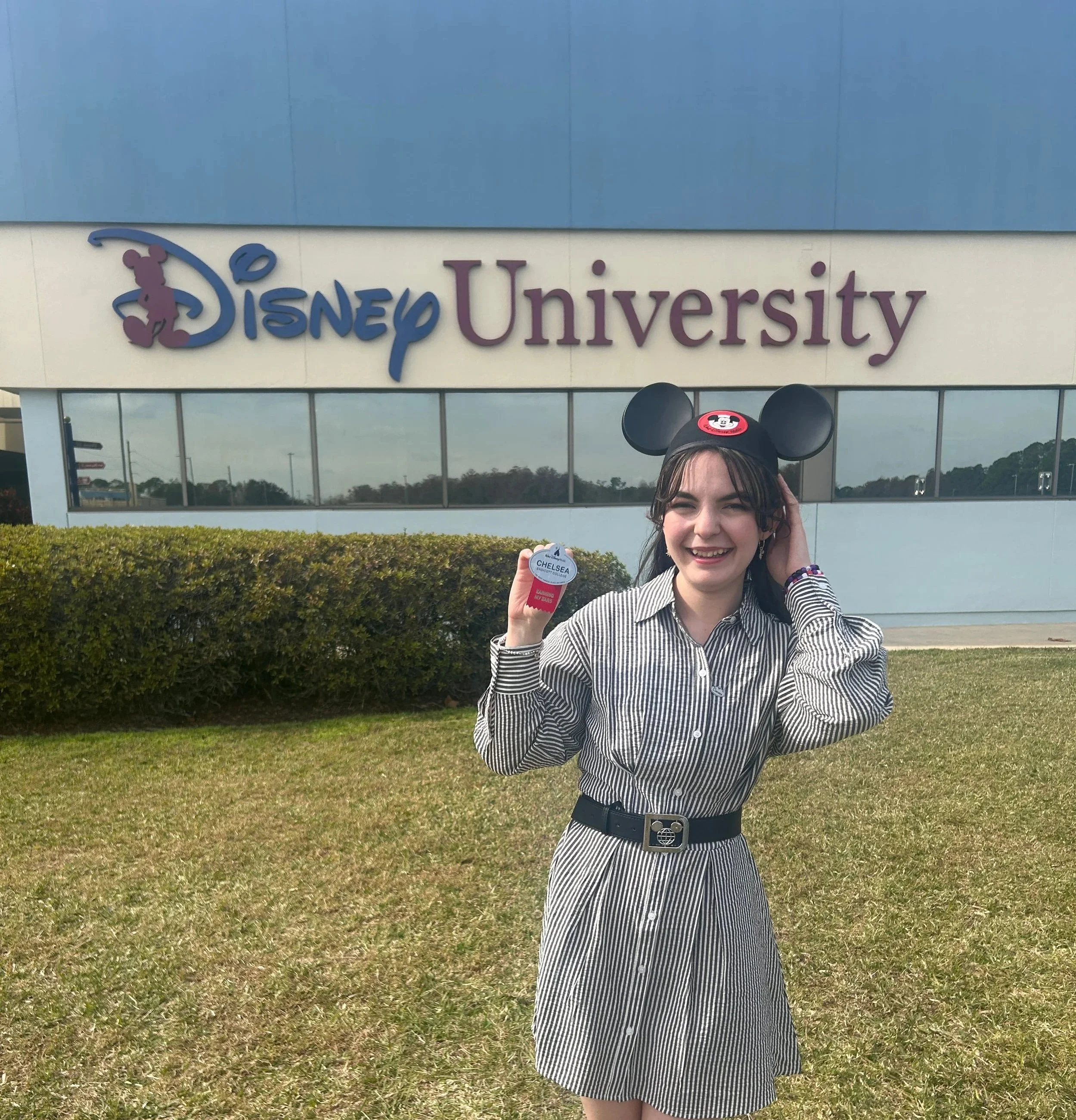 Chelsea poses in front of Disney University wearing a classic Mickey hat and holding her Cast Member name tag.