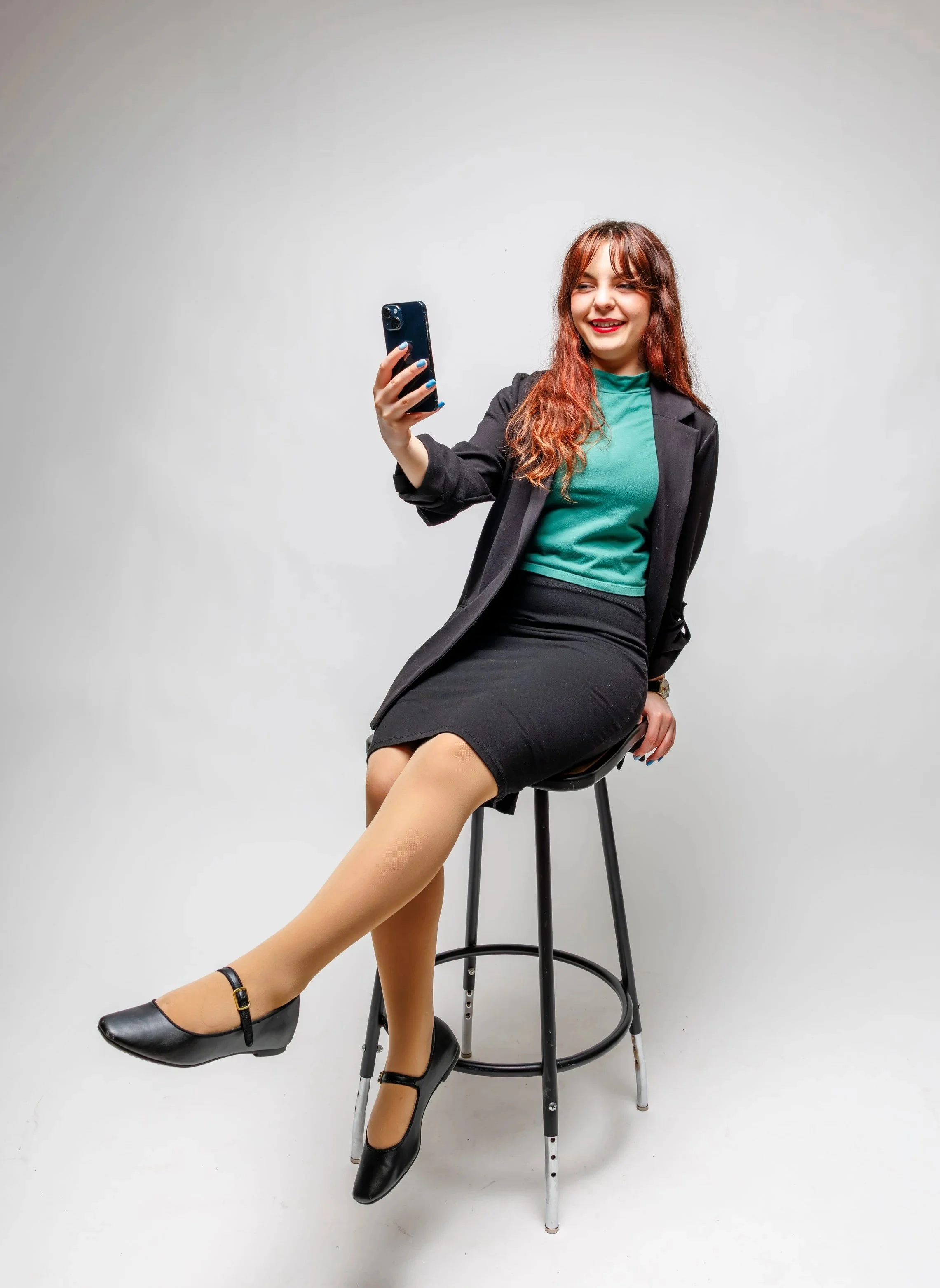 Chelsea, in professional dress, sits on a stool smiling at her iPhone.