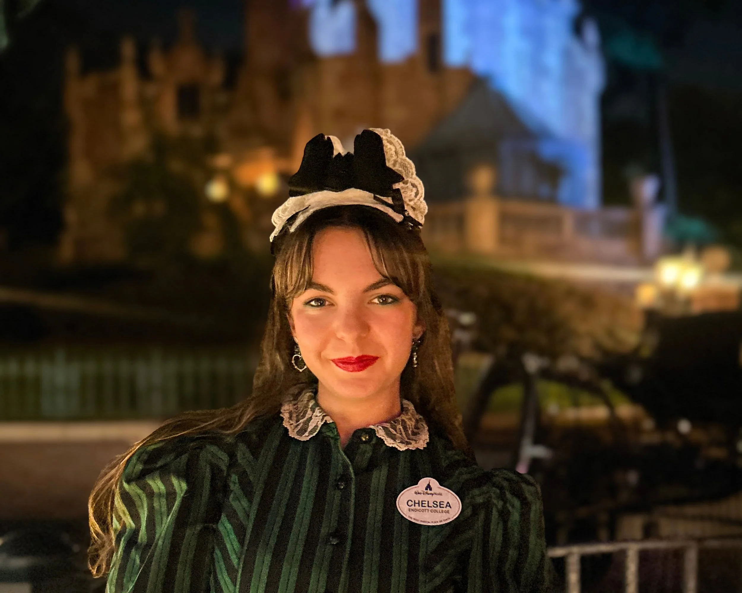 Chelsea, dressed in her Cast Member costume, is standing in front of the Haunted Mansion at night.