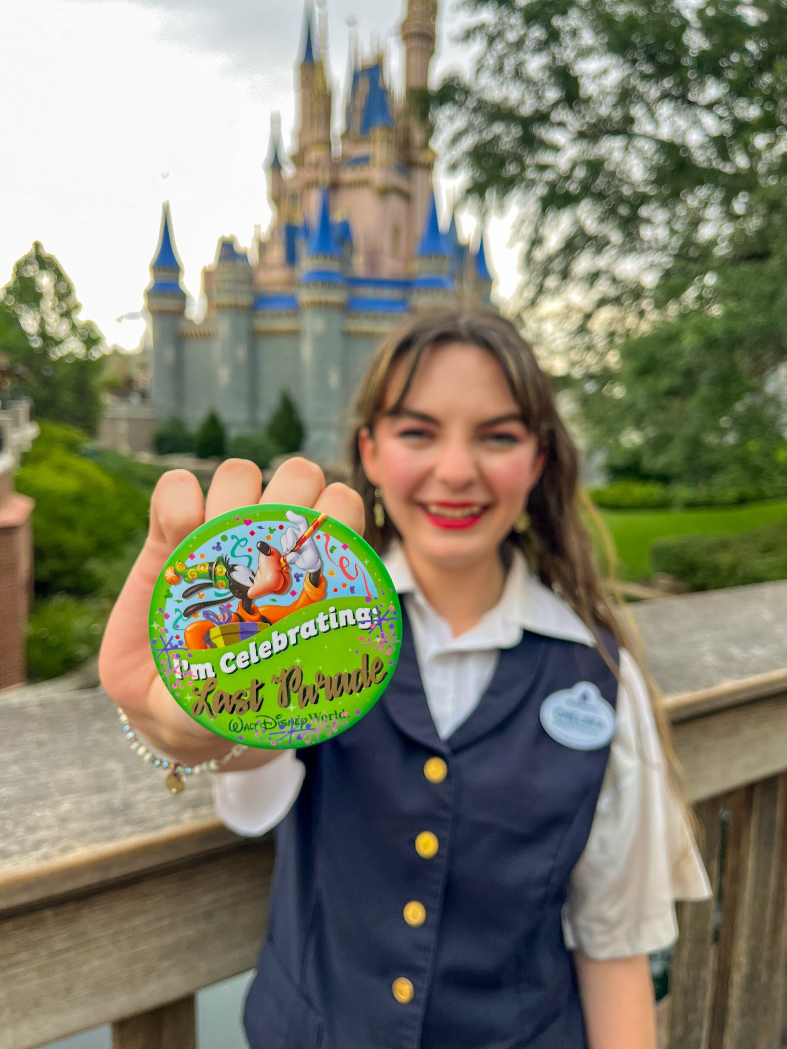Chelsea poses in front of Cinderella Castle in her Guest Flow Team Cast Member costume holding a celebration button that says "Last Parade".