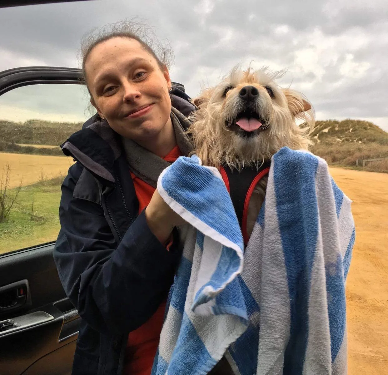 A woman drying wet muddy paws holding a happy, small, fluffy dog wrapped in a blue and white towel outdoors on a cloudy day.