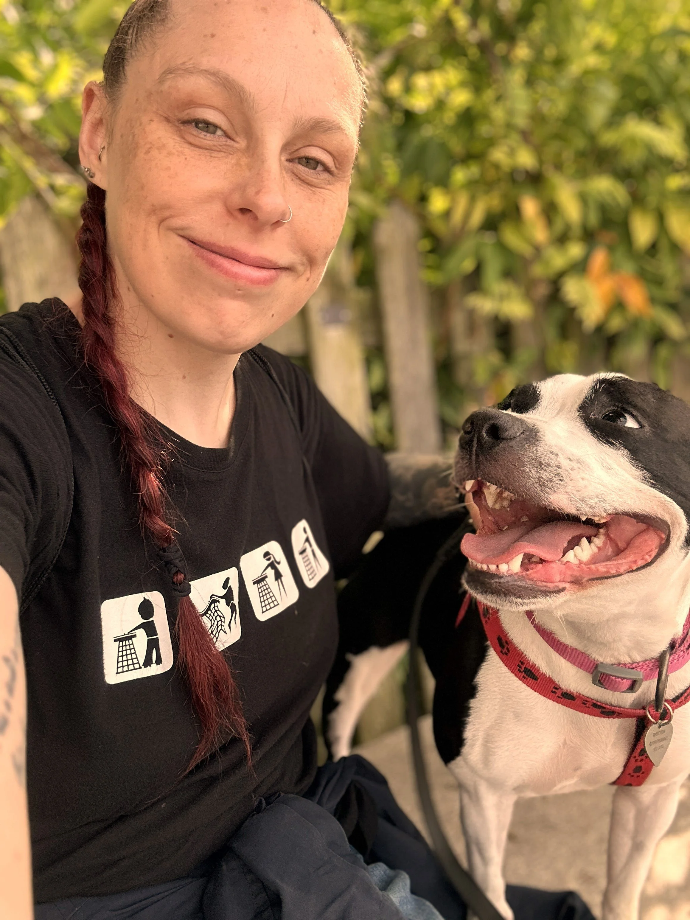 A woman with a braid smiling while sitting next to a happy black and white dog with a red collar in a park or garden setting.