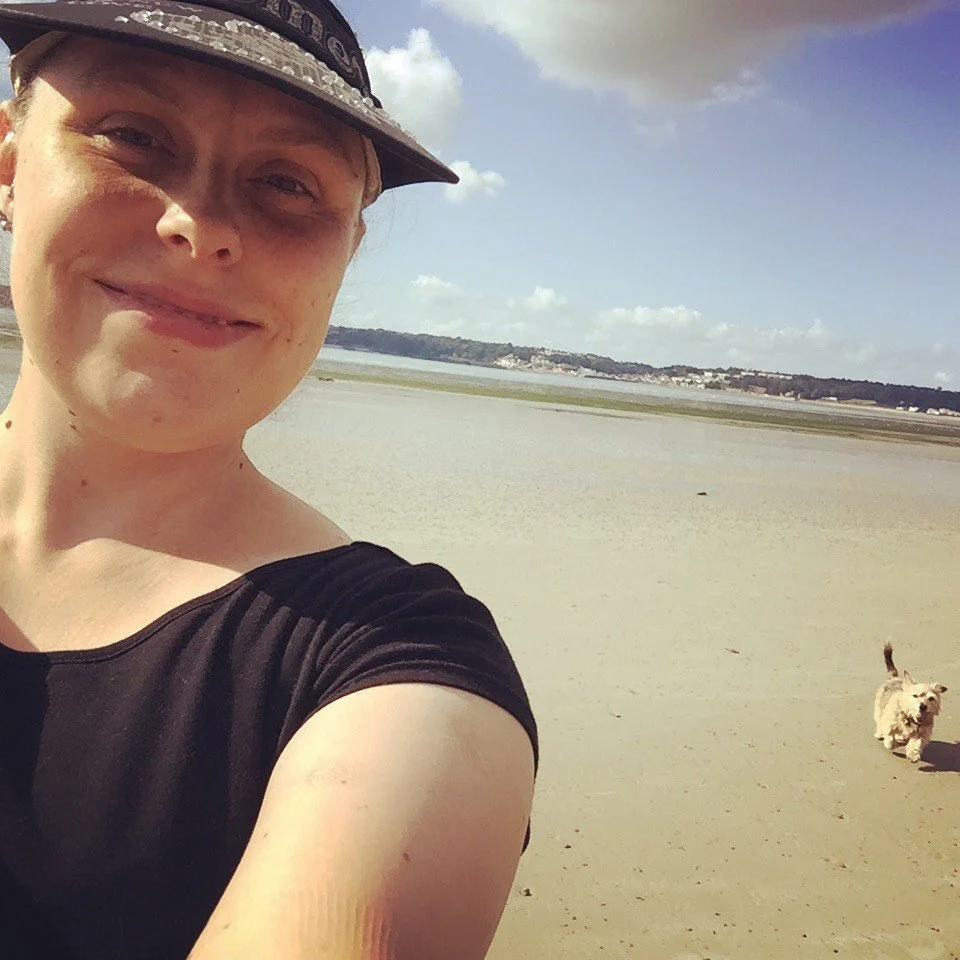 A woman smiling at the camera taking a selfie on a sandy beach, with a small dog running in the background and water and clouds in the distance.
