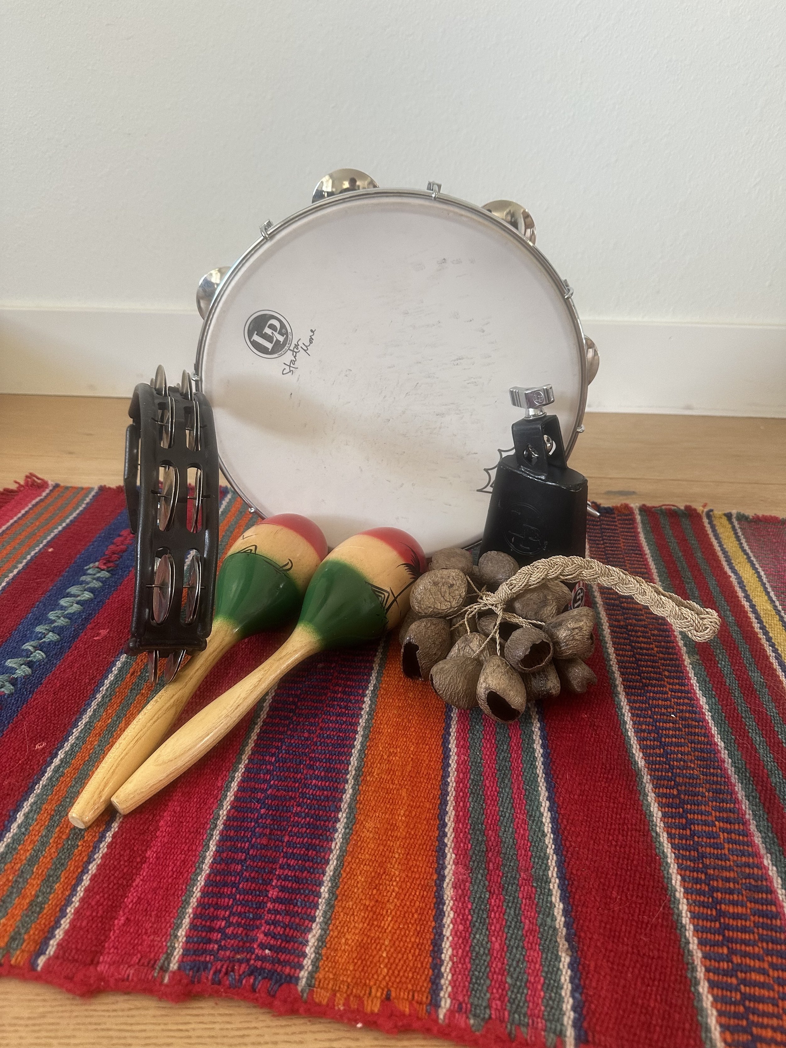 A collection of musical instruments including a tambourine, a pair of maracas, a recorder, and a cluster of small dried seed pods on a colorful woven cloth.