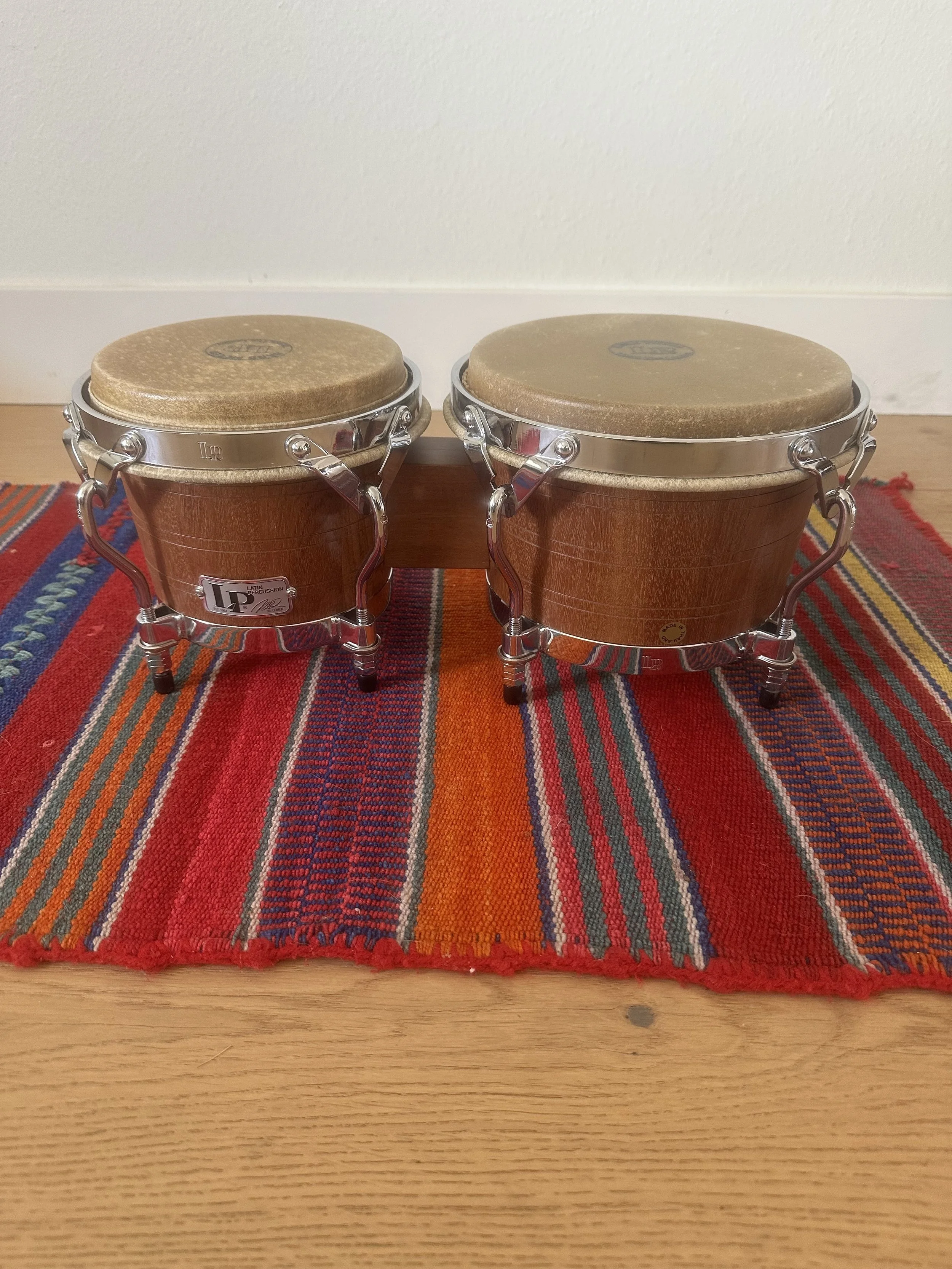Pair of bongo drums with wooden shells, metal rims, and drumheads, placed on a red, orange, and blue woven rug on a wooden floor against a white wall.