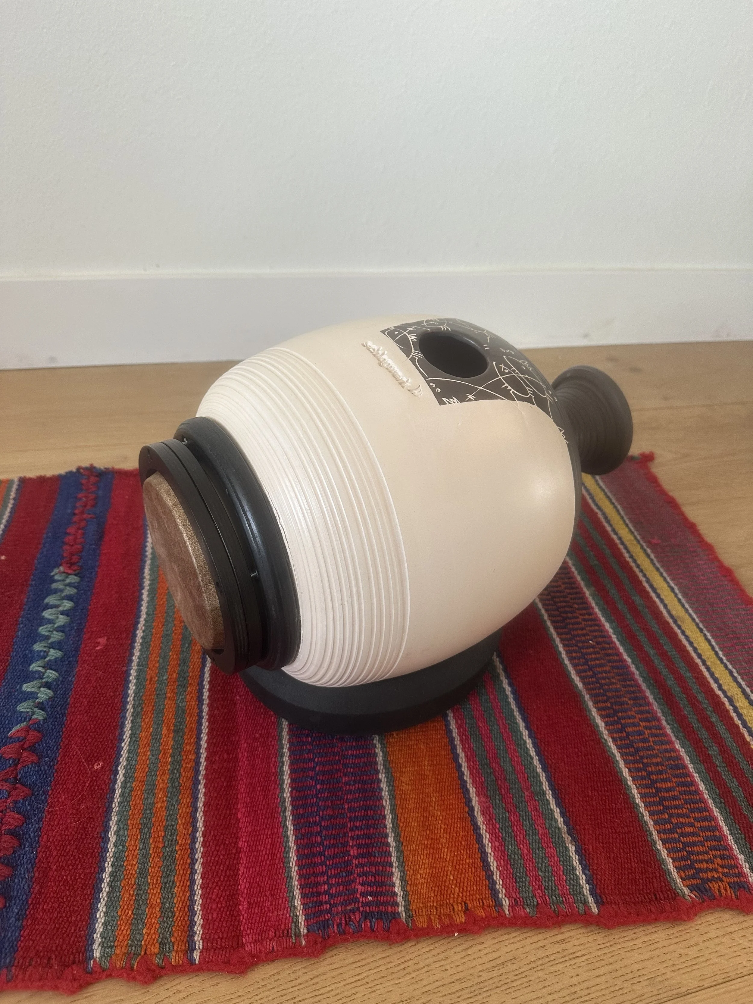 A teapot-shaped object with a black handle and spout, resting on a colorful striped cloth on a wooden floor.