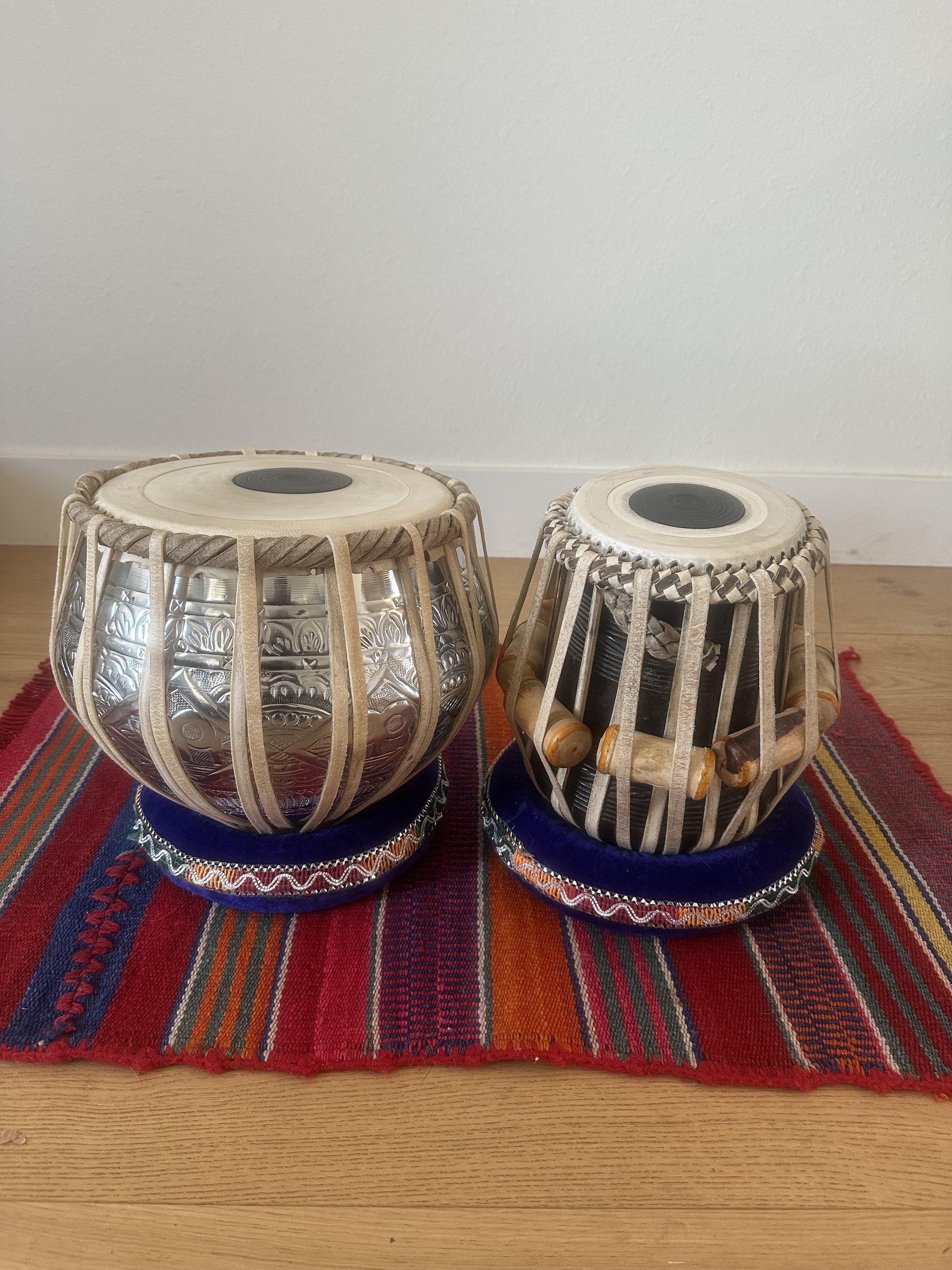 Two traditional Indian tabla drums with decorative cushions on a striped red, orange, and blue cloth on a wooden surface.