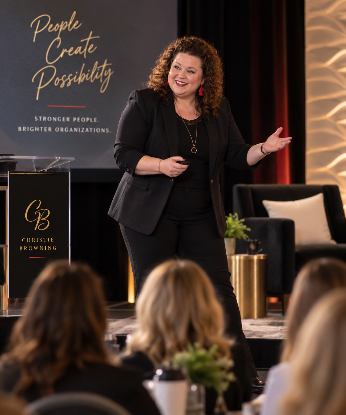 A woman with curly hair giving a presentation on stage, wearing a black blazer and pants, with a large screen behind her displaying the text "People Create Possibility" and "Stronger People, Brighter Organizations." She appears engaged and smiling, with audience members in the foreground.