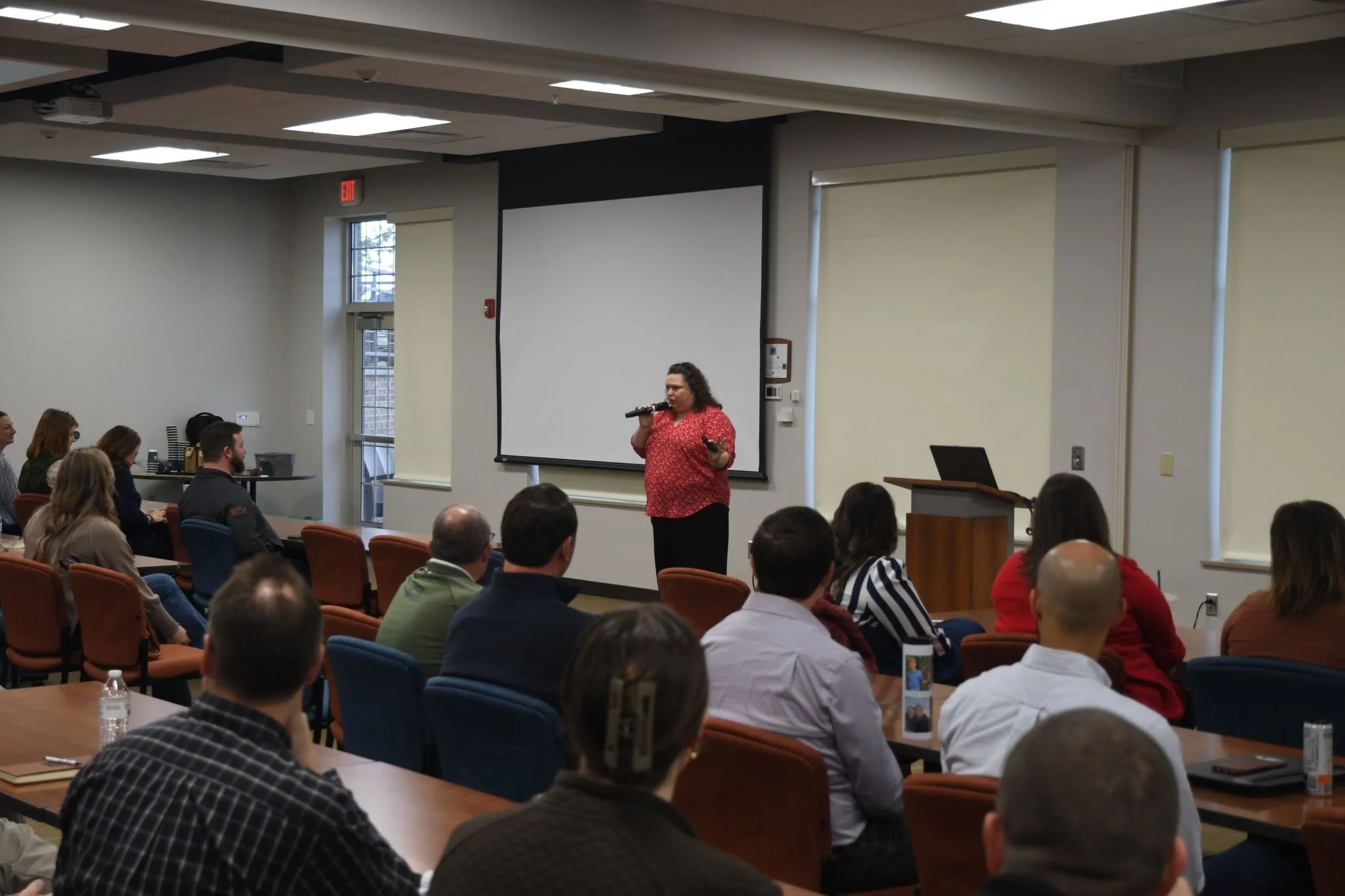 A woman in a red patterned shirt holding a microphone and a remote control presents to an audience in a conference room with a projection screen behind her, and several people seated listening.