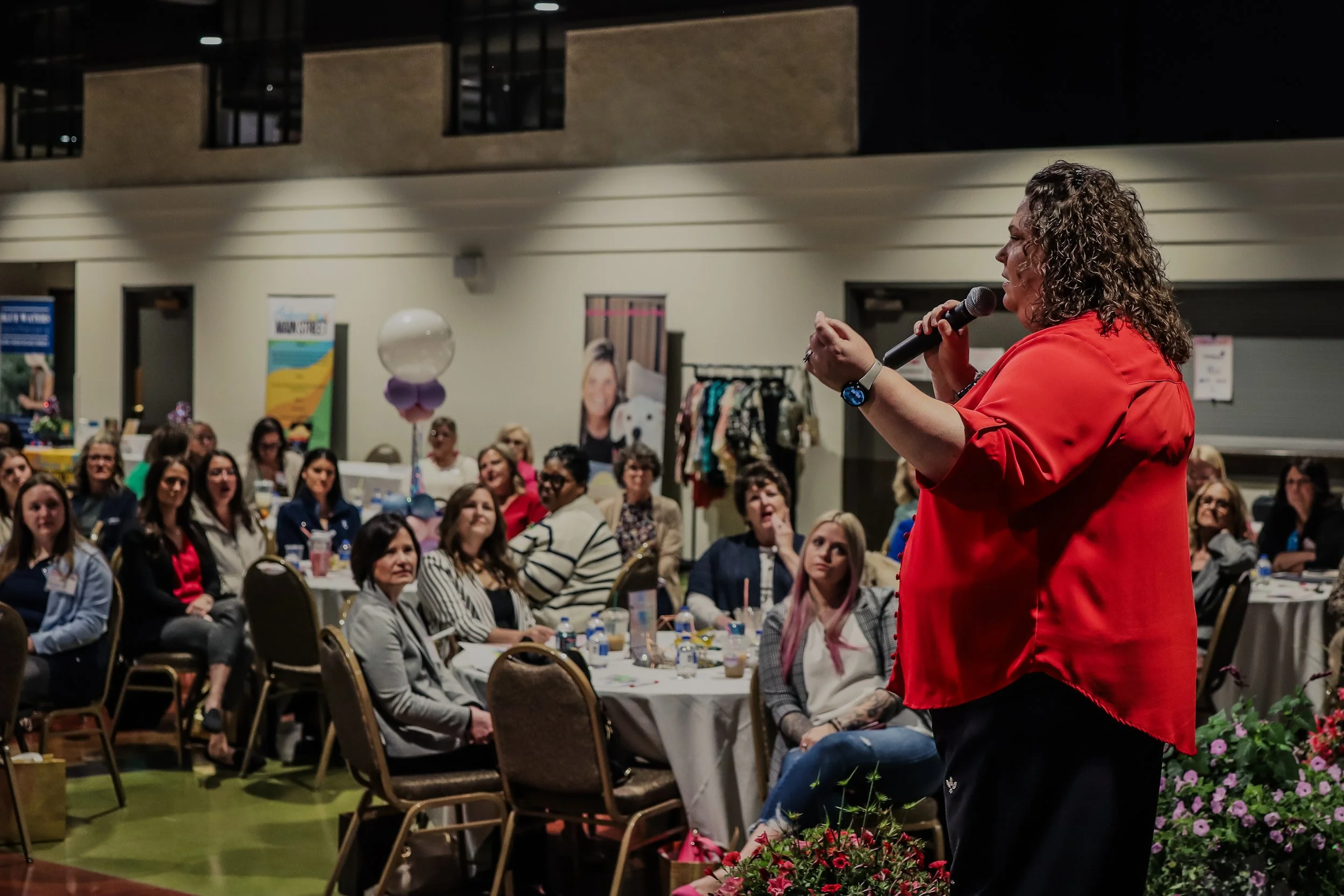 A woman with curly hair wearing a red blazer speaking into a microphone at a conference or seminar, with an audience of diverse women listening attentively, seated at round tables decorated with flowers and balloons in an indoor venue.
