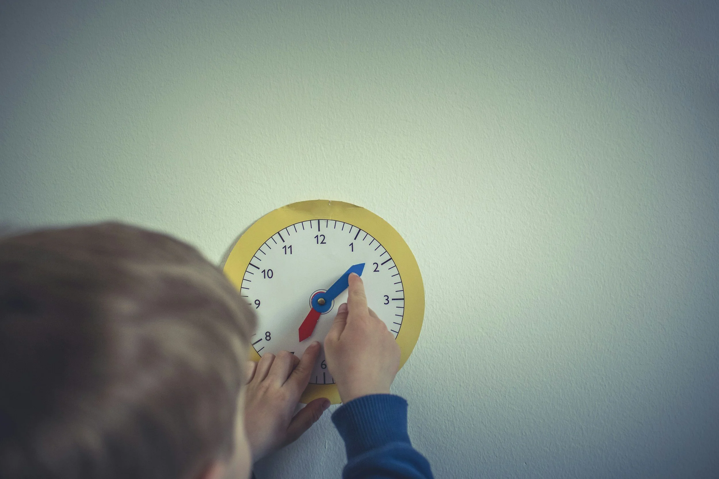 Child adjusting a yellow clock on a light green wall, indicating the time as 1:10.