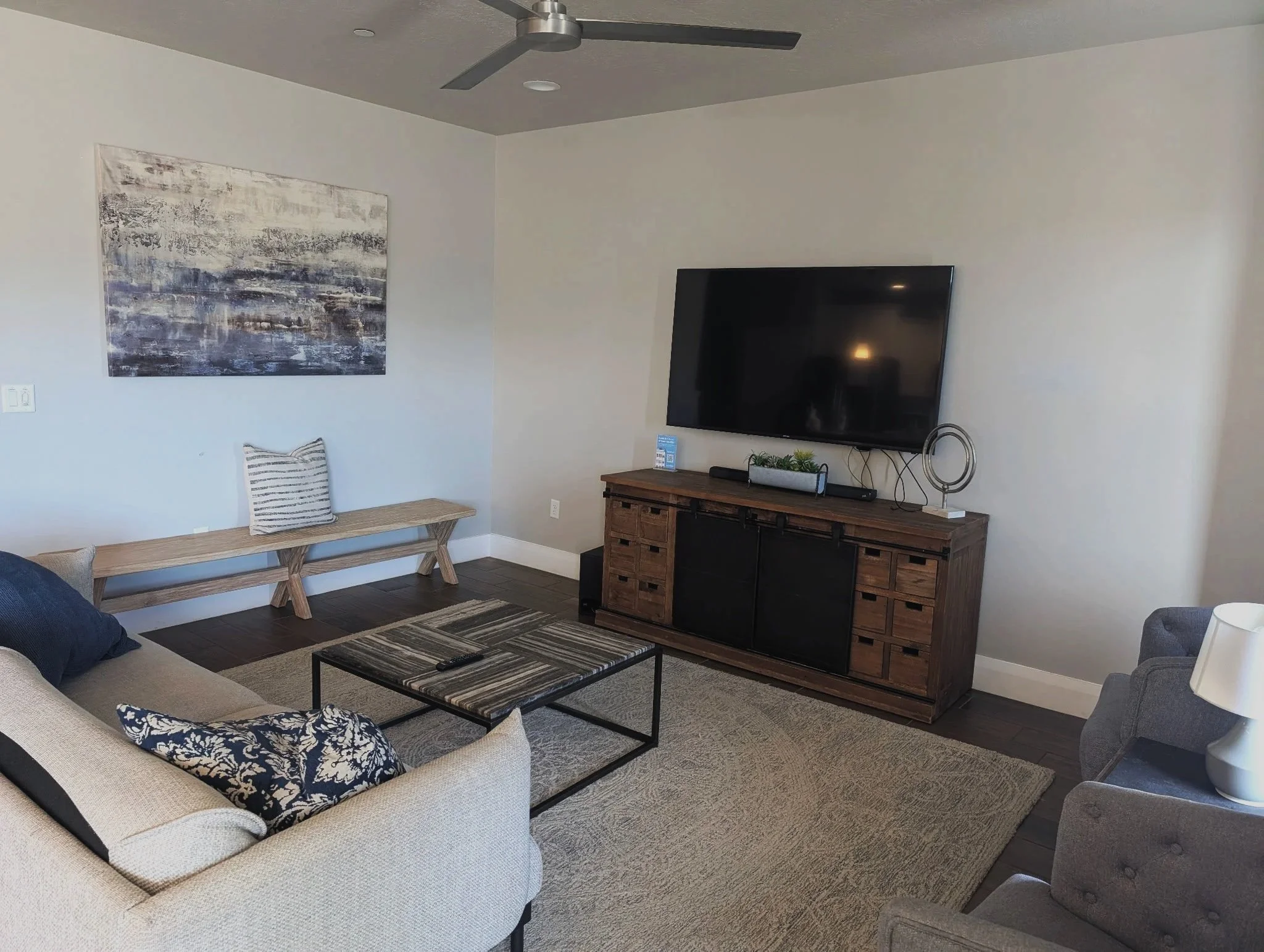 Living room with a beige sofa, a gray armchair, a wooden TV stand with a flat-screen TV, a coffee table, and wall art.