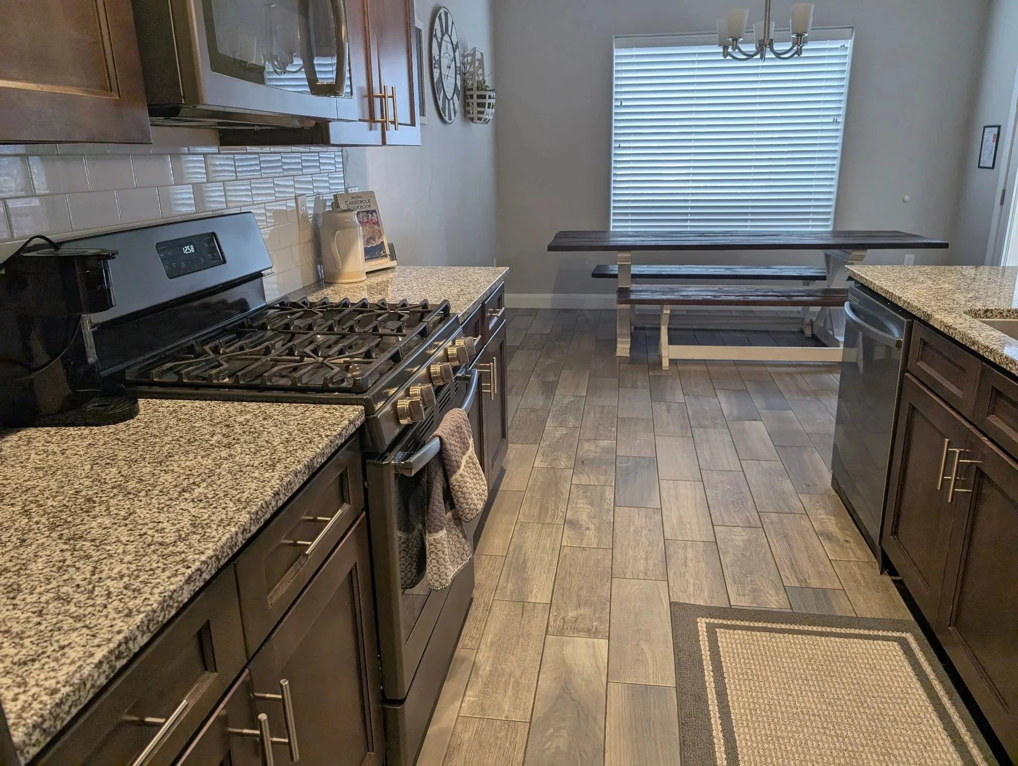 Clean kitchen with dark wood cabinets, granite countertops, stainless steel appliances, a coffee maker, and a dining table at the back with a chandelier and window blinds.
