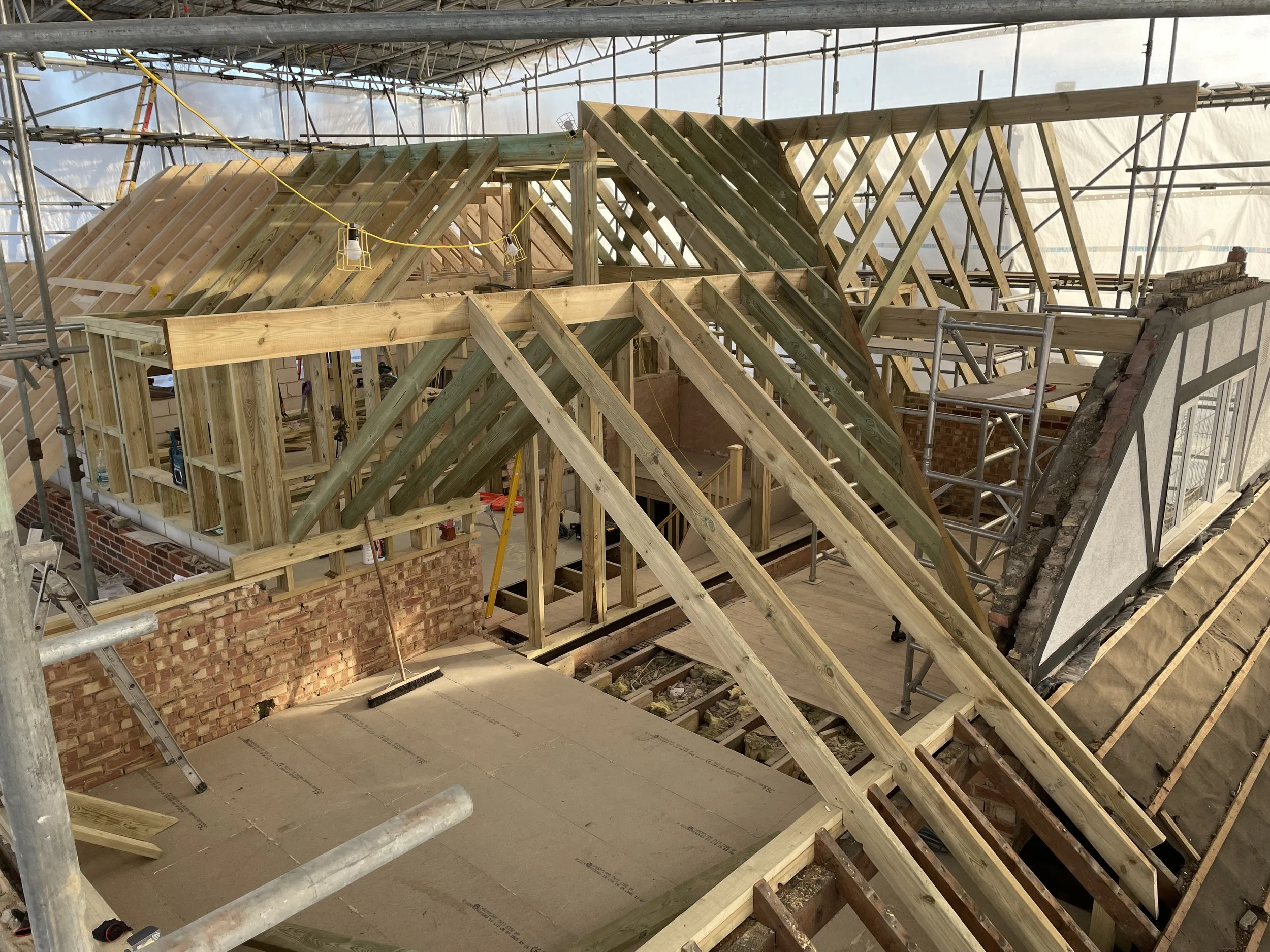 Construction site with wooden framing for a roof or upper floor, scaffolding surrounds the area, and a brick wall is partially built.