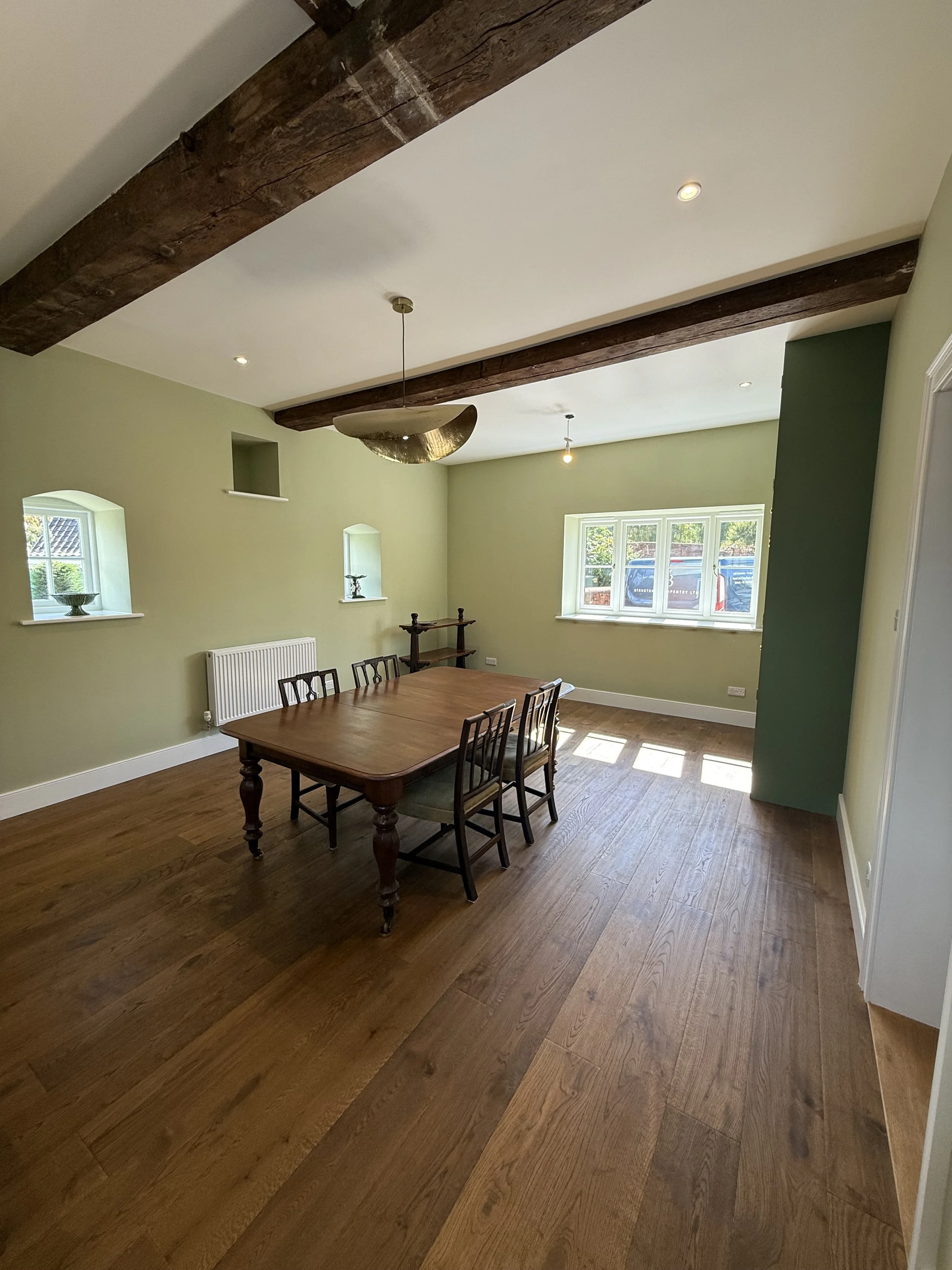 A dining room with a wooden table and chairs, green walls, windows with sunlight, and wooden ceiling beams.