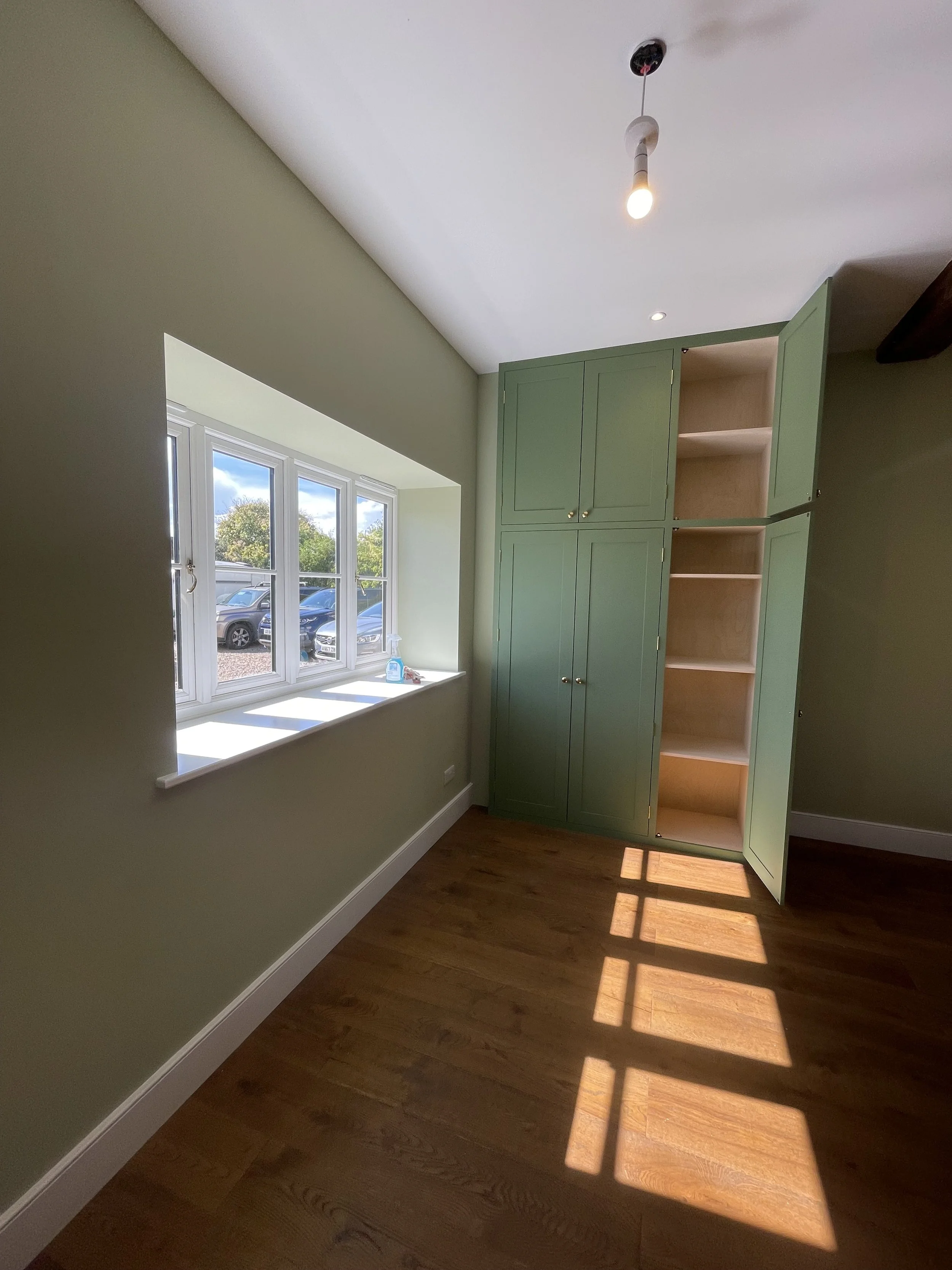 Room with green built-in cabinet, window with sunlight casting shadows on wooden floor, ceiling pendant light, and outside parking lot visible through the window.