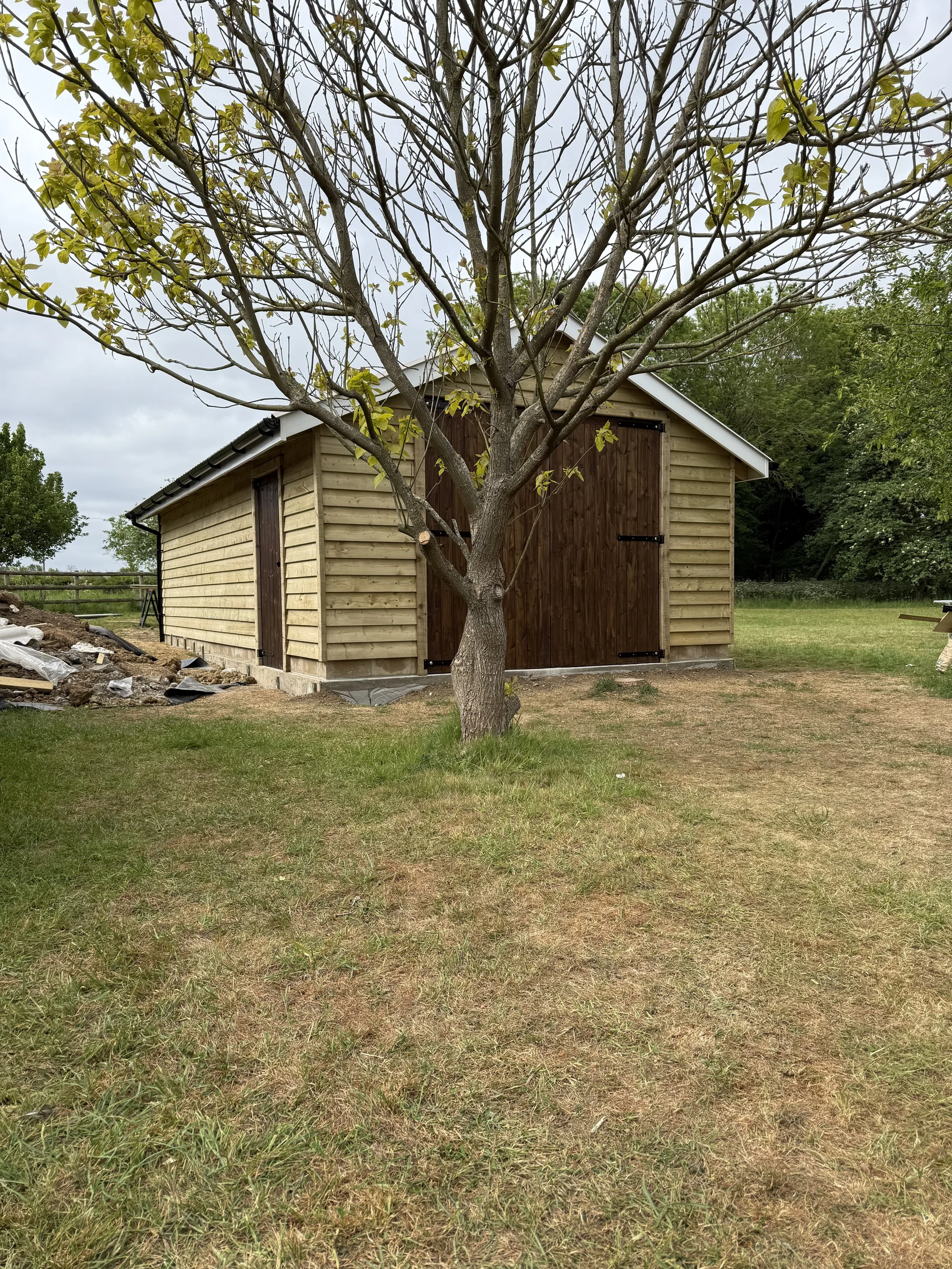 A small wooden shed with a sloped roof and closed double door, situated in a grassy yard, with a leafless tree in front of the shed and green trees in the background under an overcast sky.