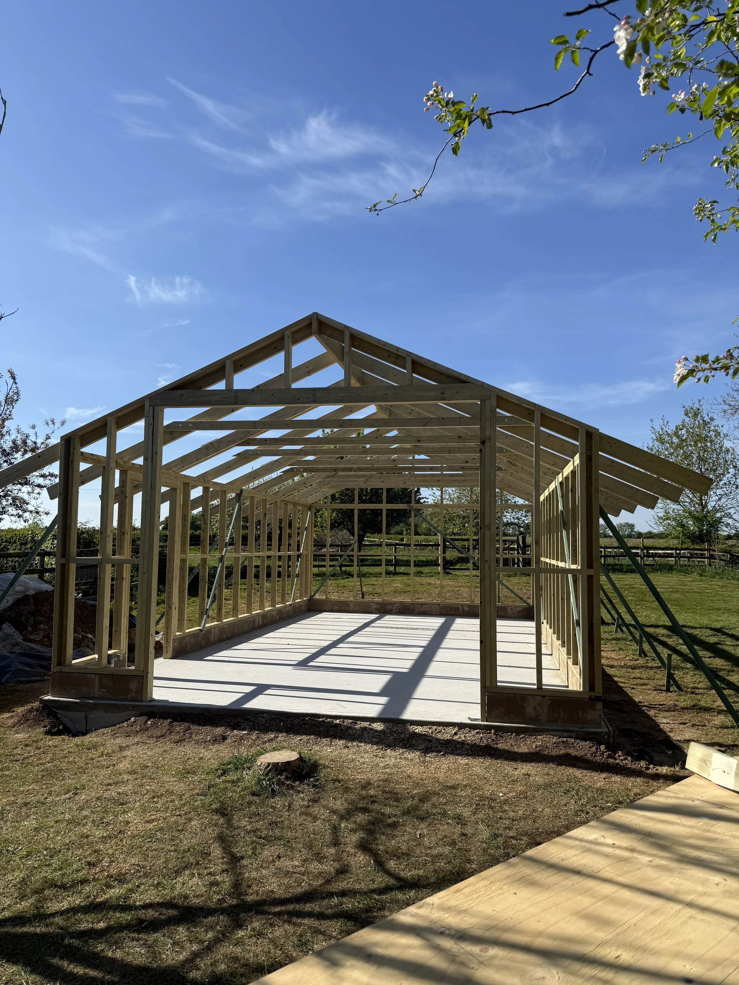 Wooden house frame under a clear blue sky, surrounded by trees and grass.