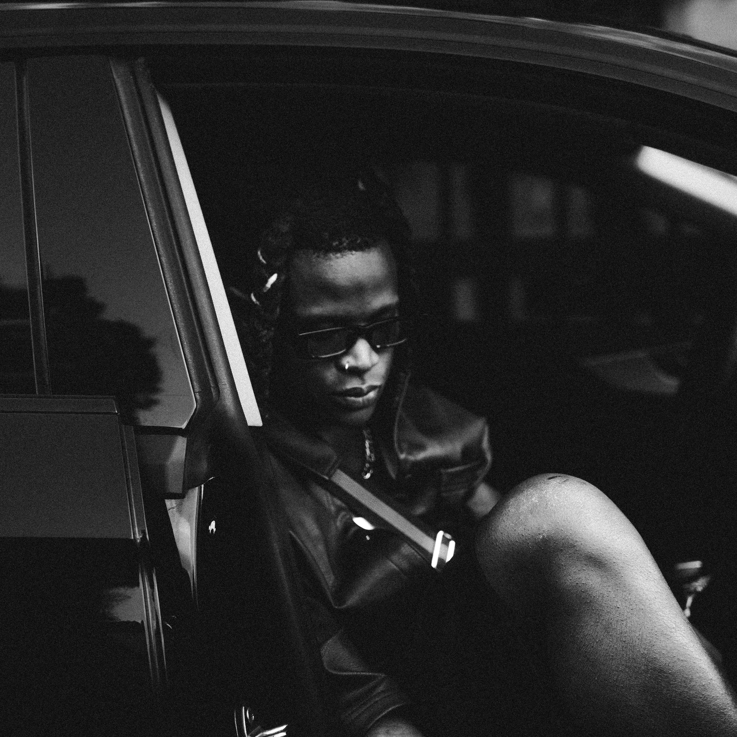 A woman with dreadlocks and sunglasses sitting in a car, wearing a leather jacket, black and white photo.