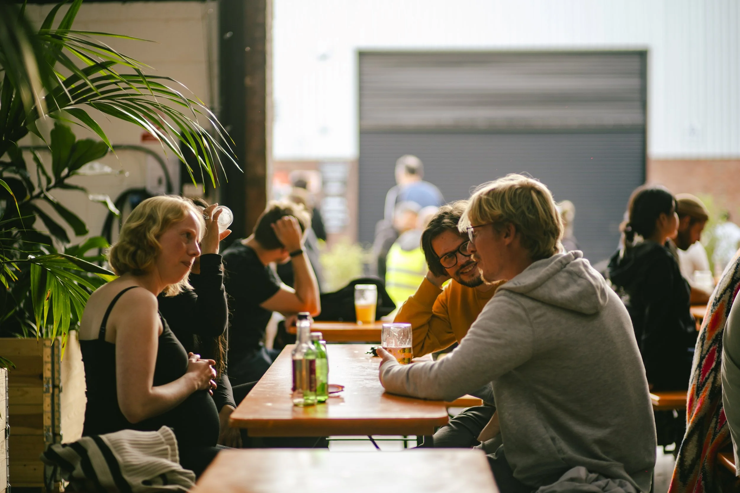 People sitting at a bar or brewery, socializing and drinking beer, with some engaged in conversation, in a bright, industrial-style setting with plants and a large open garage door.