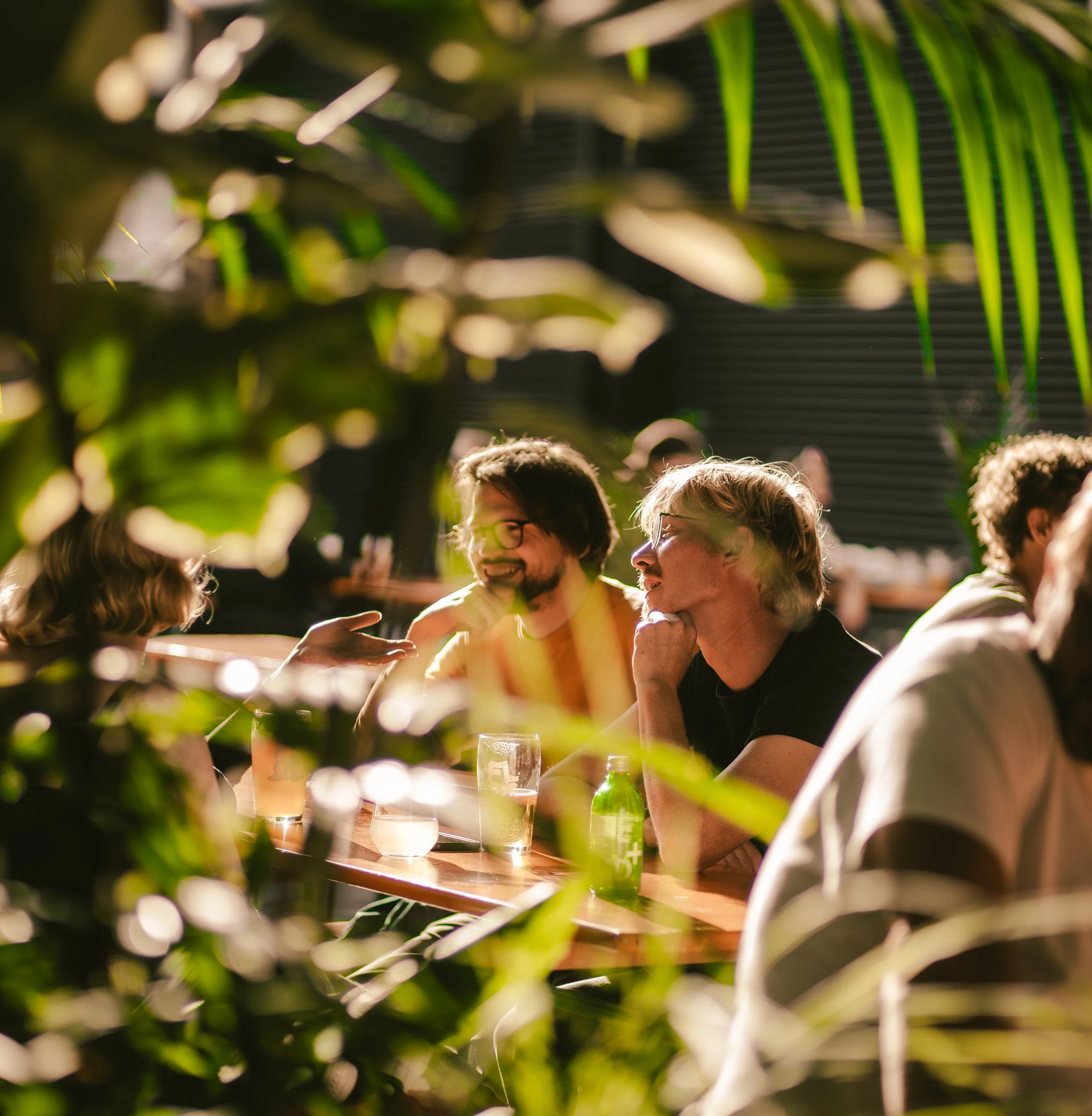 People sitting at a table in a plant-filled, sunlit outdoor setting, engaging in conversation and smiling.