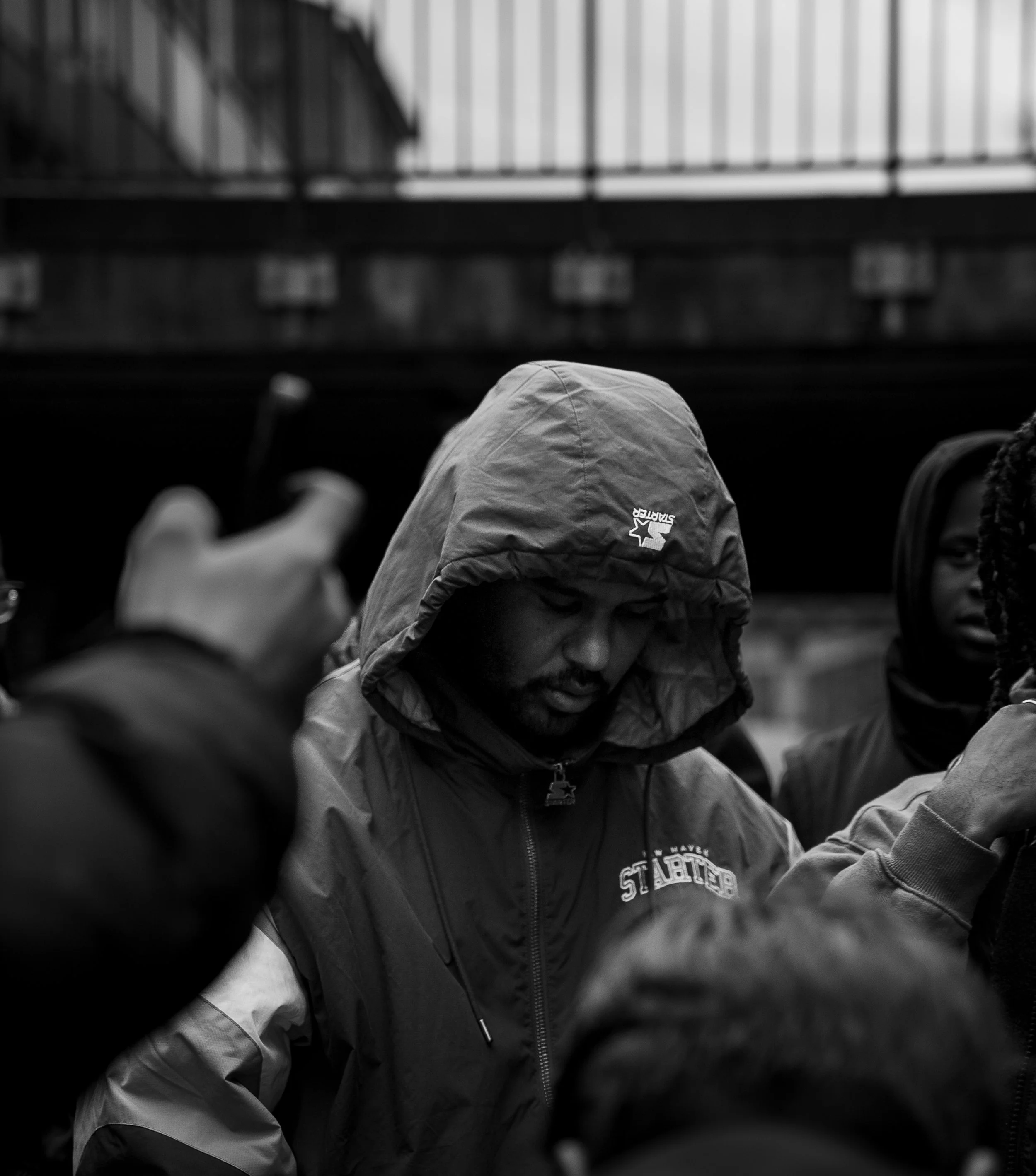 A black and white photo of a man with a hood over his head, wearing a jacket with the word 'STARTER' visible, surrounded by several other people.