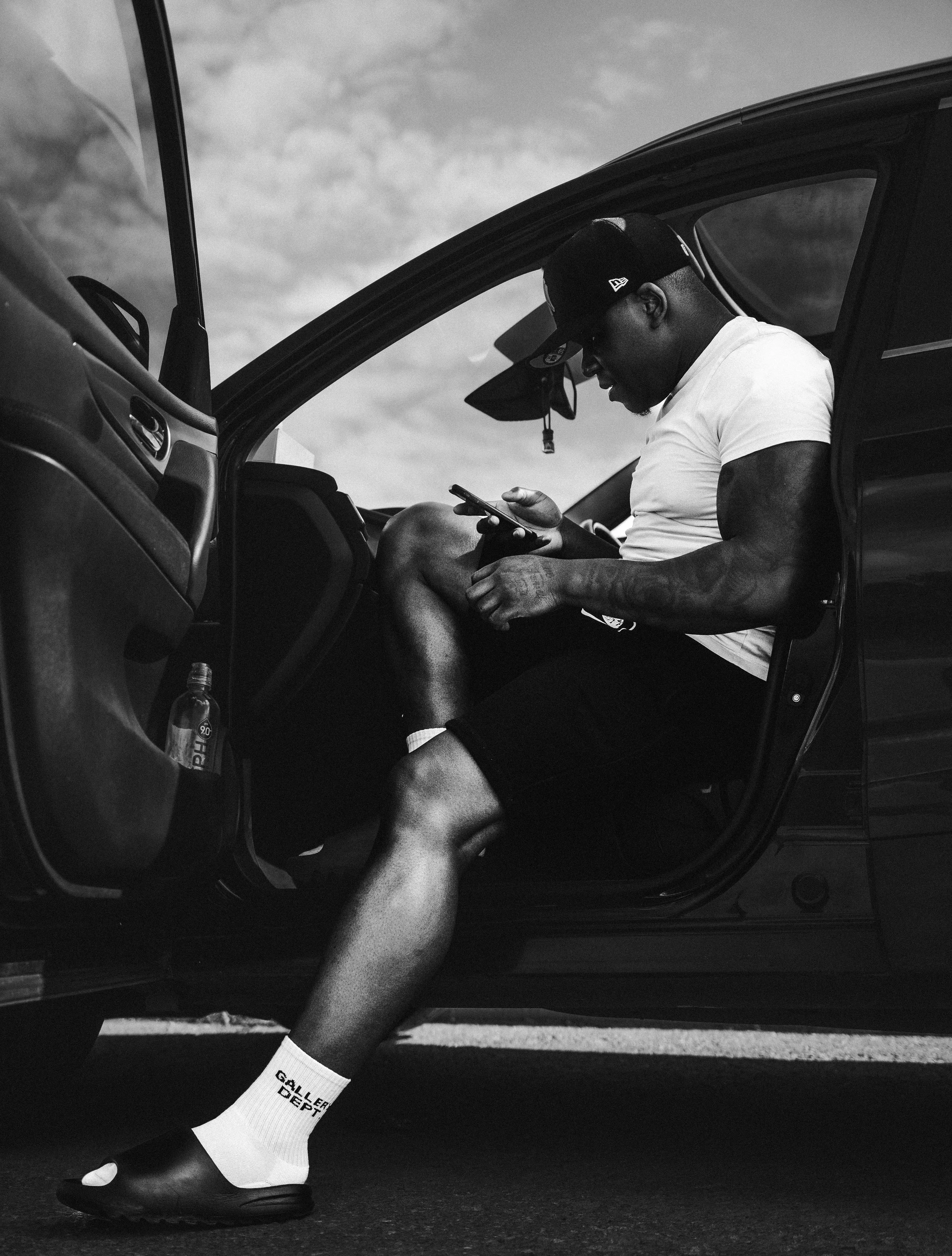 A man sitting in a car with the door open, looking at his phone, wearing a cap, t-shirt, shorts, socks, and slippers, with a water bottle in the car.