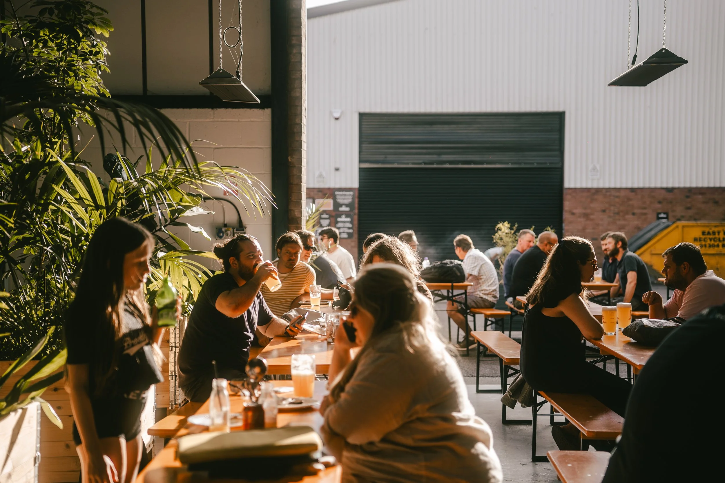 People sitting at wooden tables in a brewery or pub, enjoying drinks and socializing in a bright, industrial-style space with large open garage door and greenery.