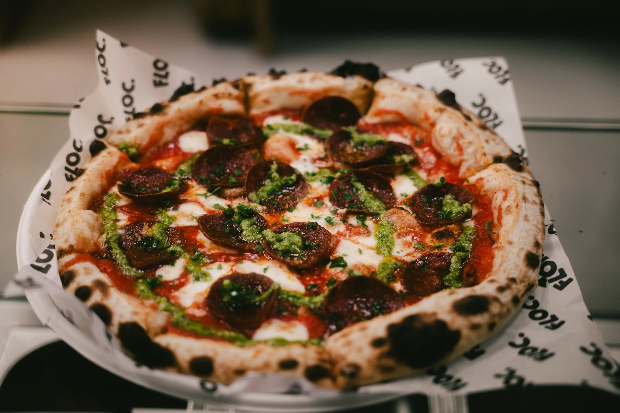 A pepperoni pizza with green pesto drizzled on top, served on a white paper-lined tray.