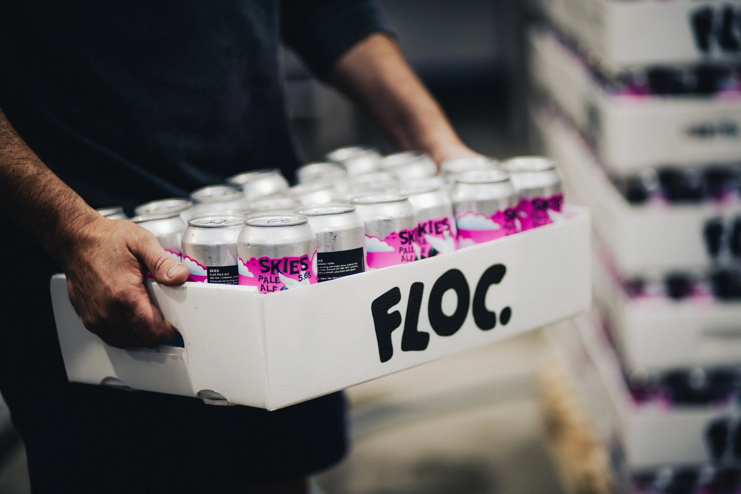 A person holding a white tray filled with cans of Skies Pale Ale beer in a store, with the word 'FLOC.' written on the side of the tray.