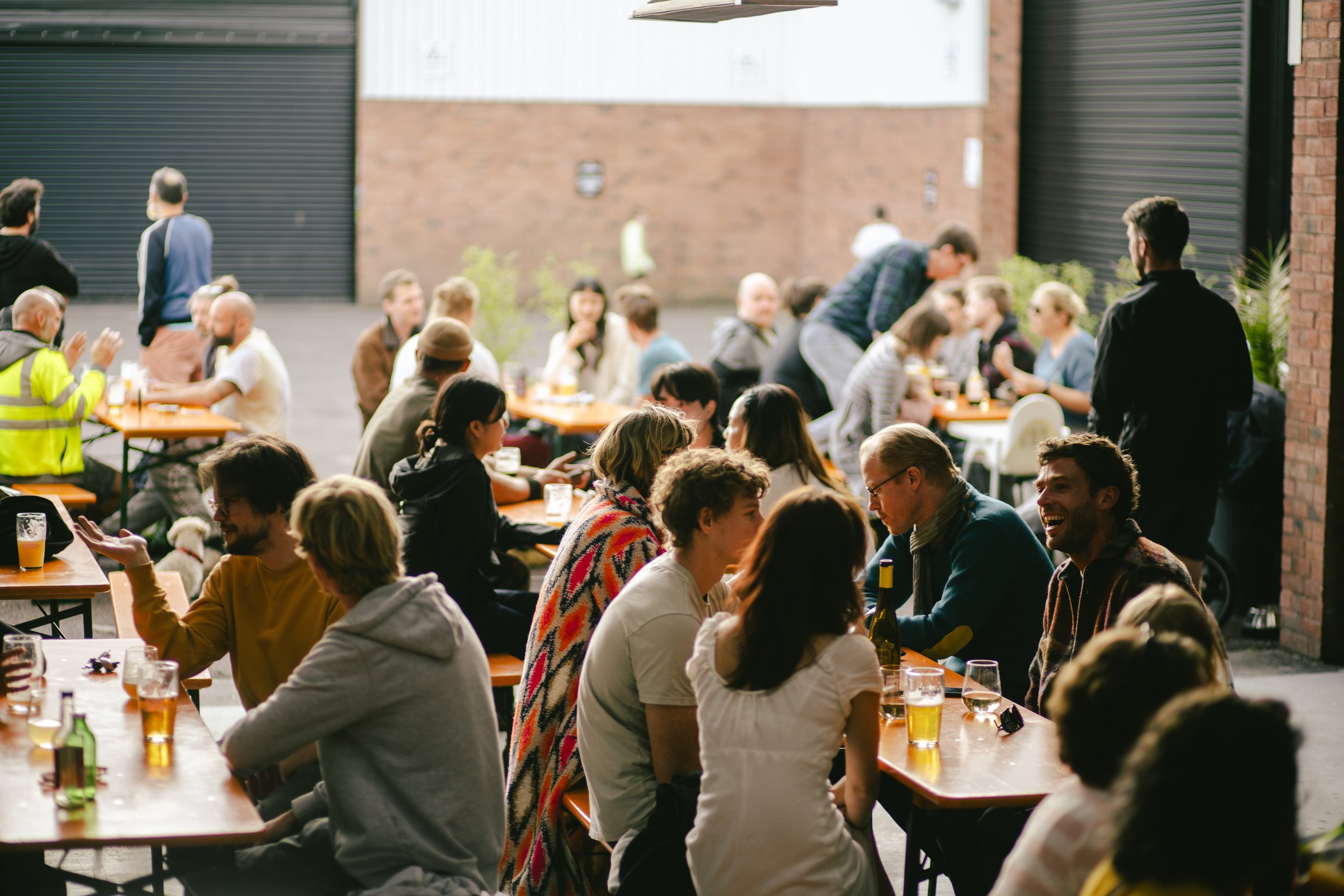 People gathered at outdoor tables in an urban setting, socializing and enjoying drinks during daytime.