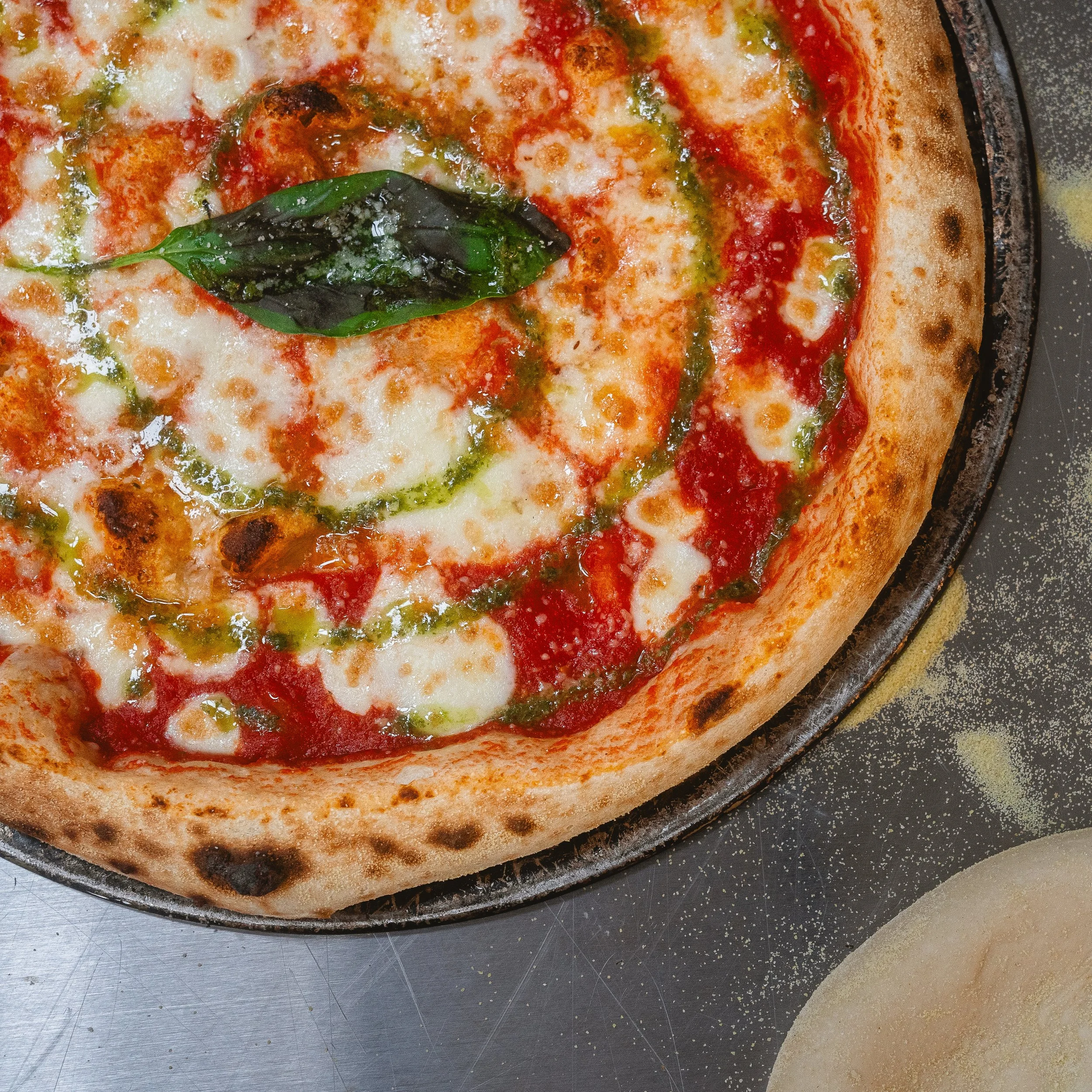 Close-up of a Margherita pizza with tomato sauce, melted mozzarella cheese, fresh basil leaf, and green pesto swirl topping, on a round metal pizza pan.