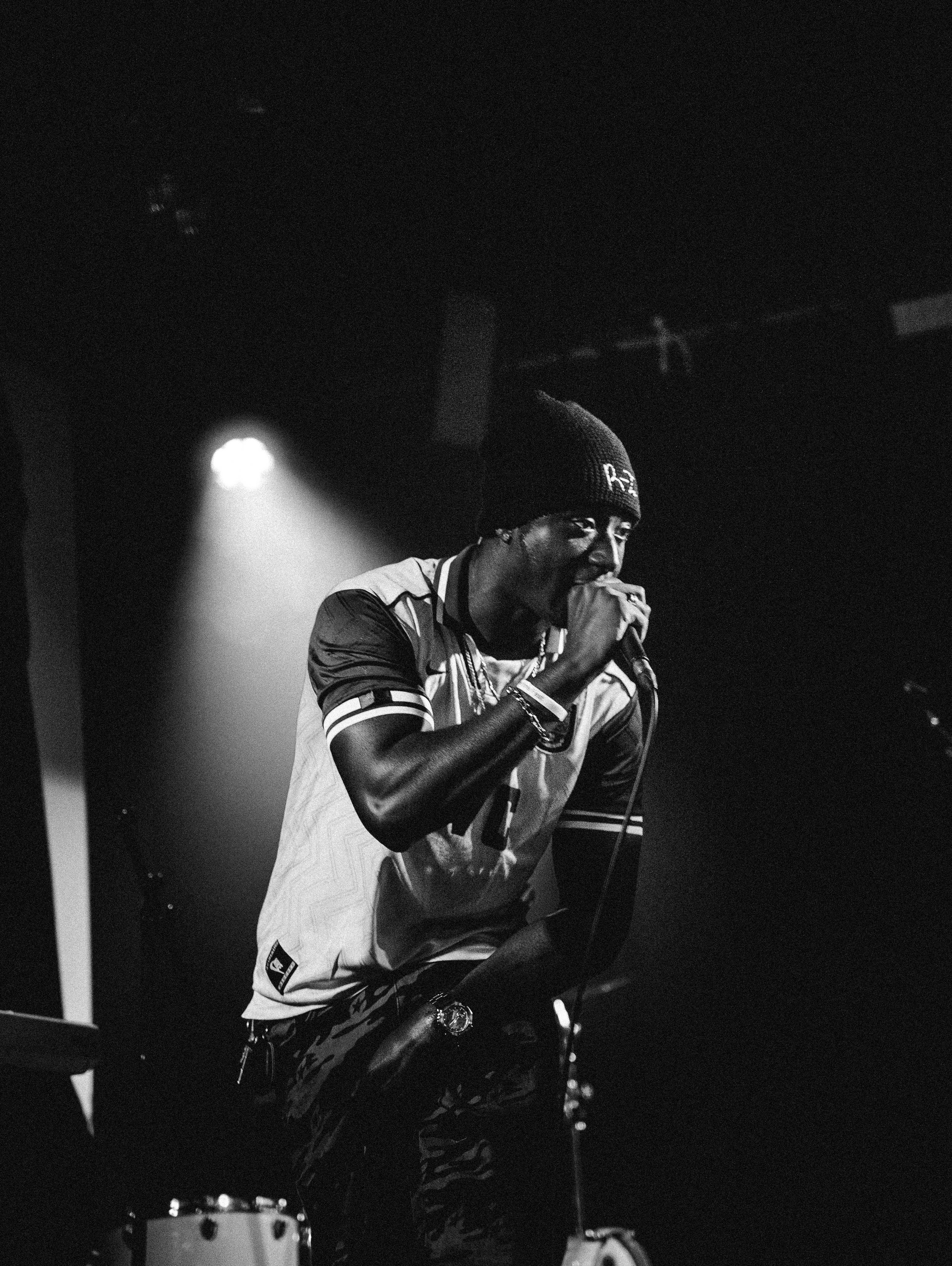 A Black man performing on stage, holding a microphone, wearing a beanie, a sports jersey, and patterned pants, with stage lights in the background.
