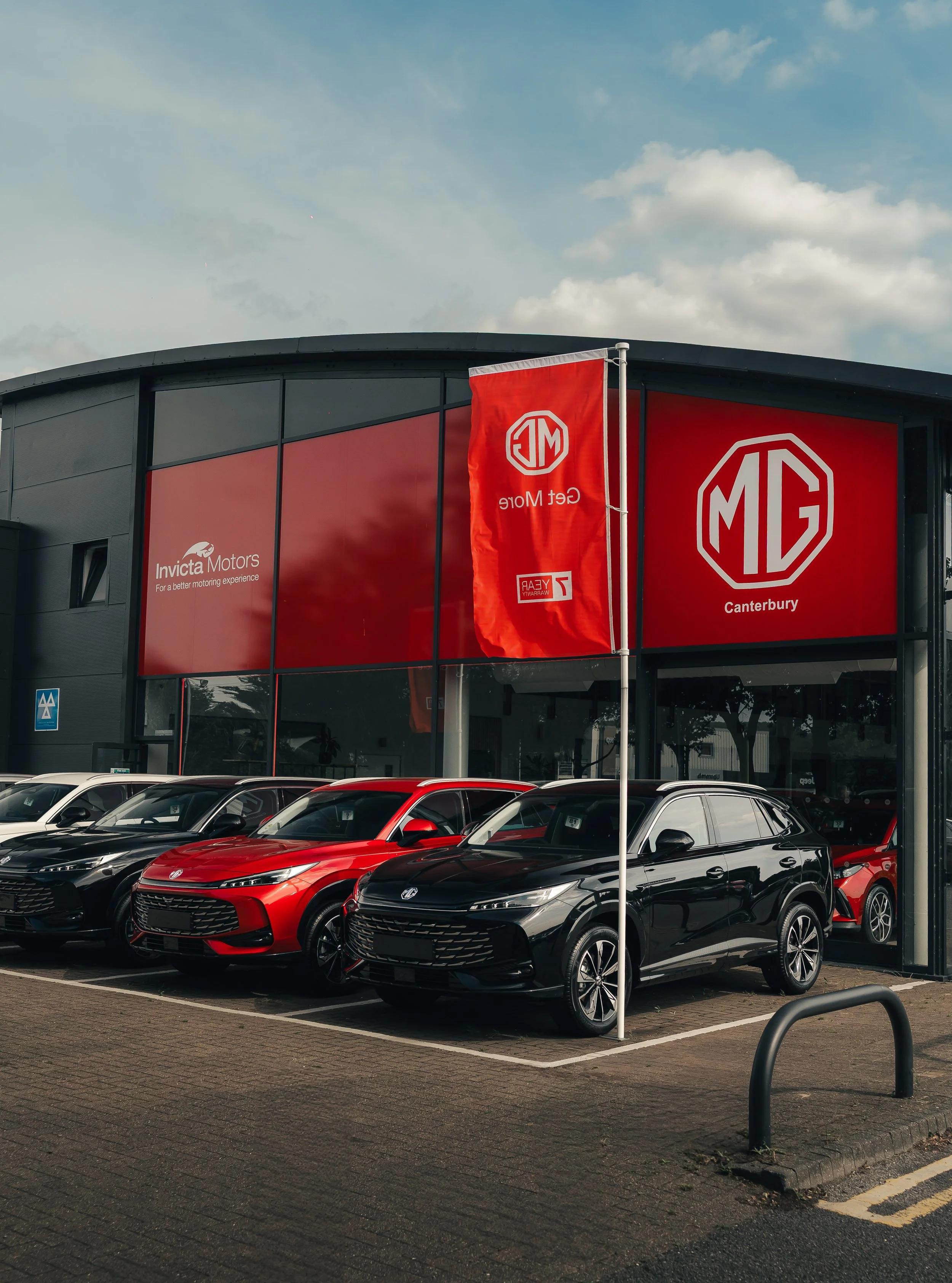 Car dealership with MG vehicles parked outside, large red MG flag, and a building with MG and Invicta Motors branding.