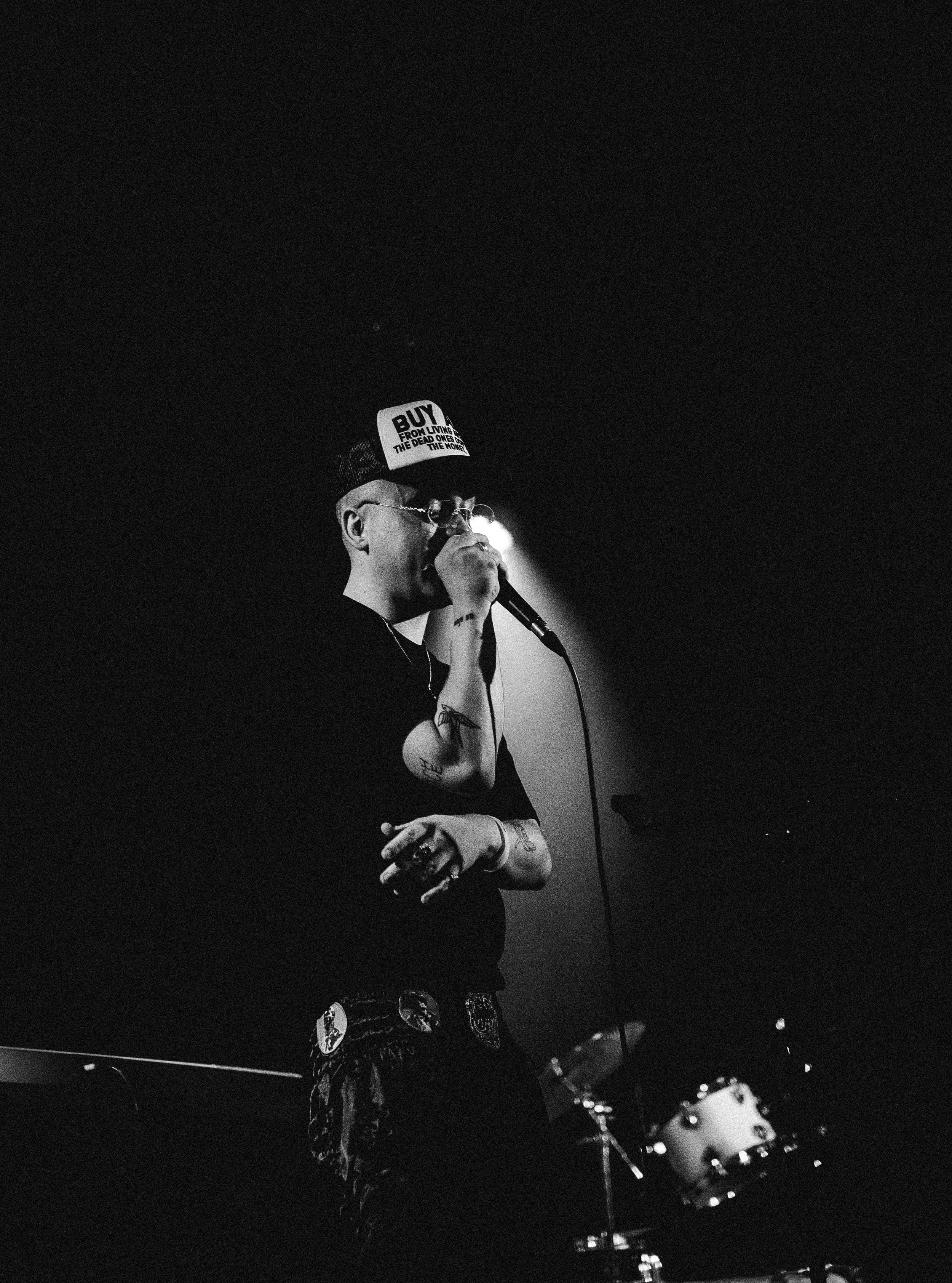 A person singing into a microphone on a dimly lit stage, wearing a hat with a sign that reads 'BUY FROM LIVING THE DEAD ONES THE NOW' and sunglasses, with tattoos on their arms, and a drum set visible in the background.