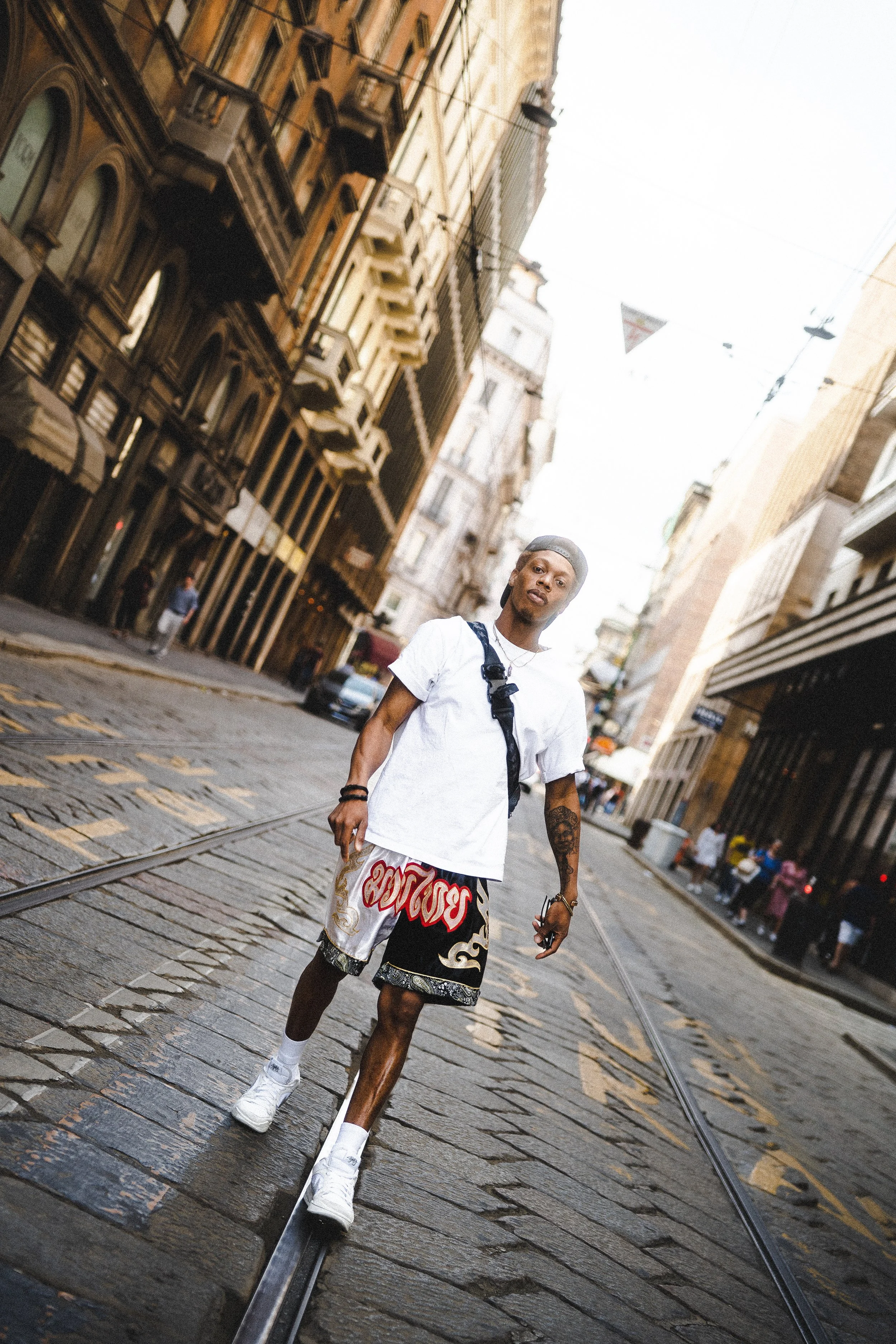 A young man walking on a city street with cobblestone pavement and tram tracks, surrounded by tall buildings.