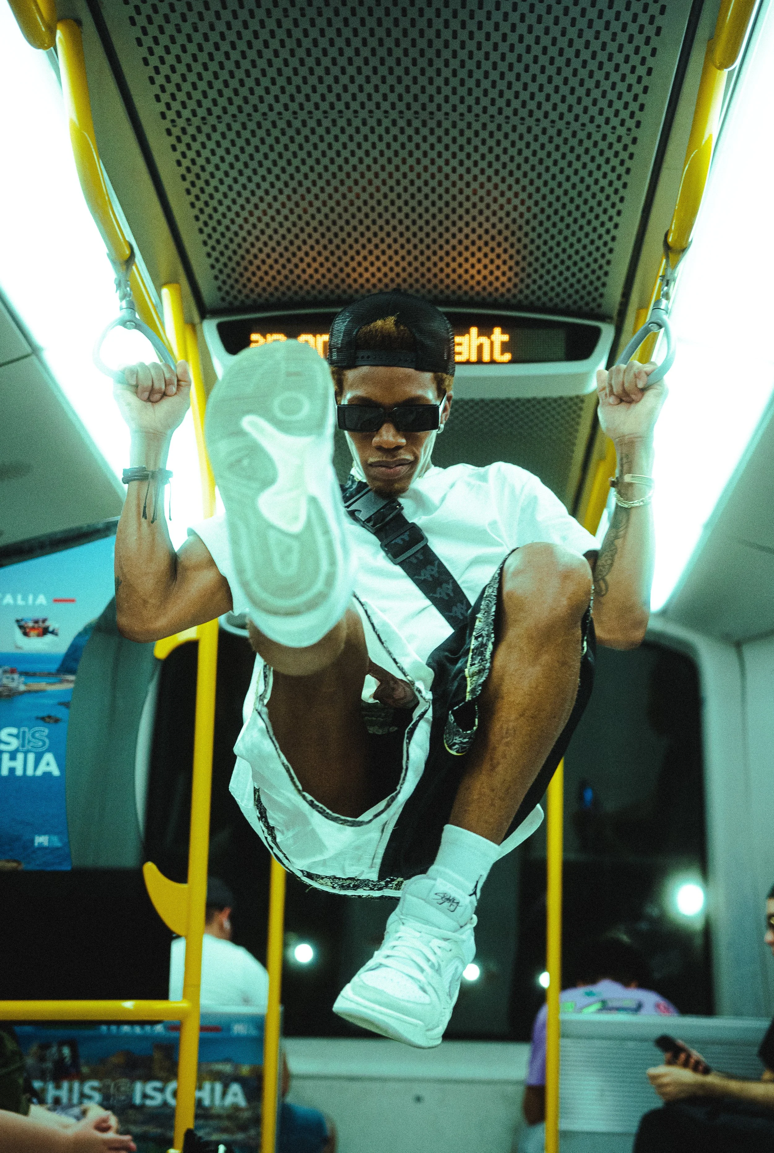 A young man wearing sunglasses, a white shirt, and a black cap rides hanging on a bus grab handle, with his leg extended forward towards the camera. The bus interior is well-lit with digital signage overhead.