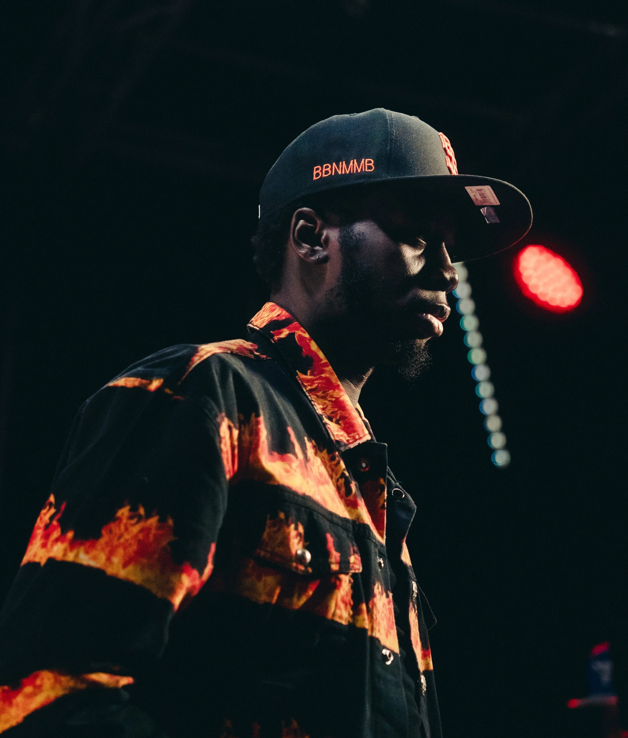 A person wearing a black baseball cap with orange text, a vibrant patterned jacket, and looking down in a dimly lit setting with colorful lights in the background.
