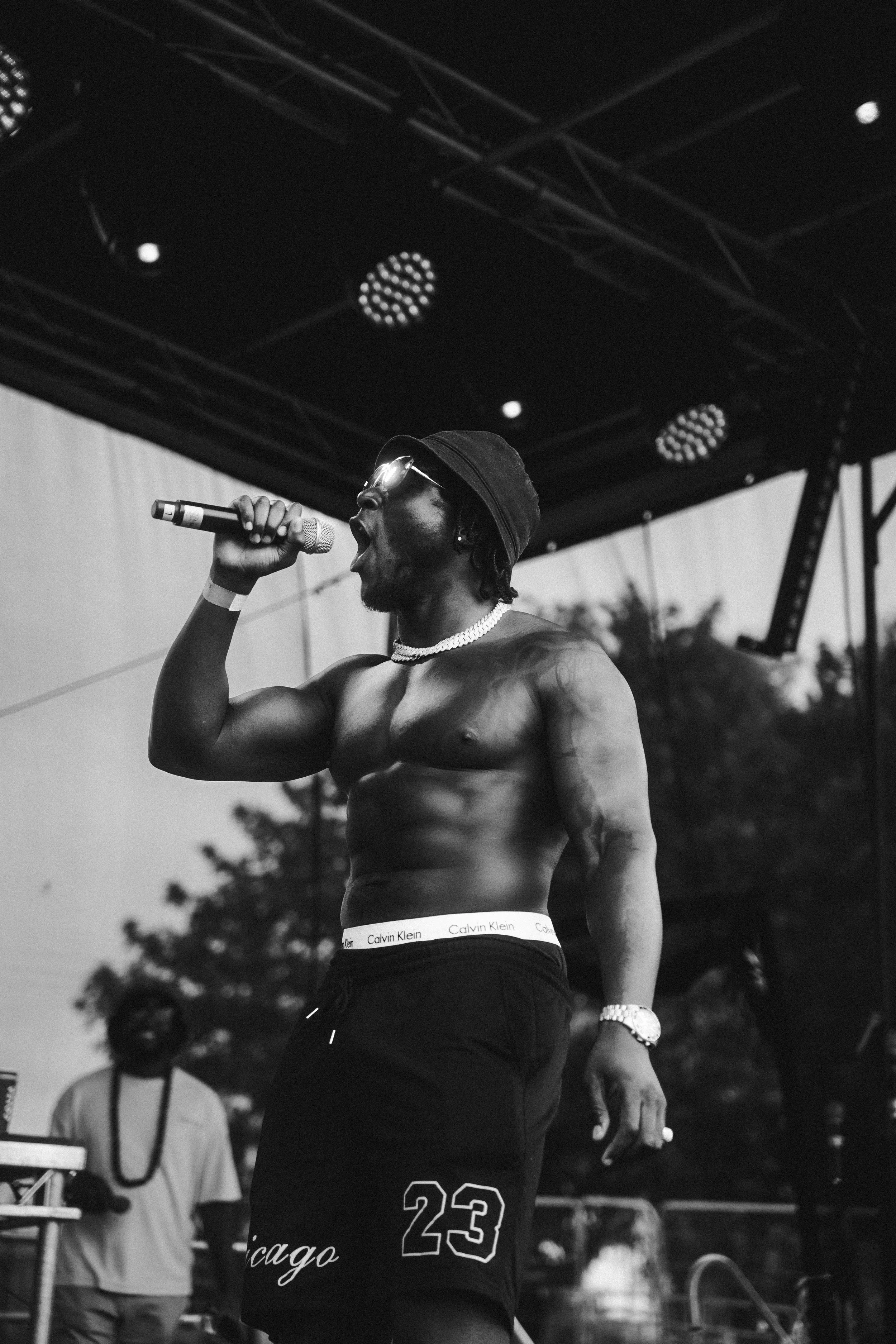 A shirtless male performer singing into a microphone on stage, wearing sunglasses, a black cap, a pearl necklace, and a wristwatch, with a dark sky and stage equipment in the background.